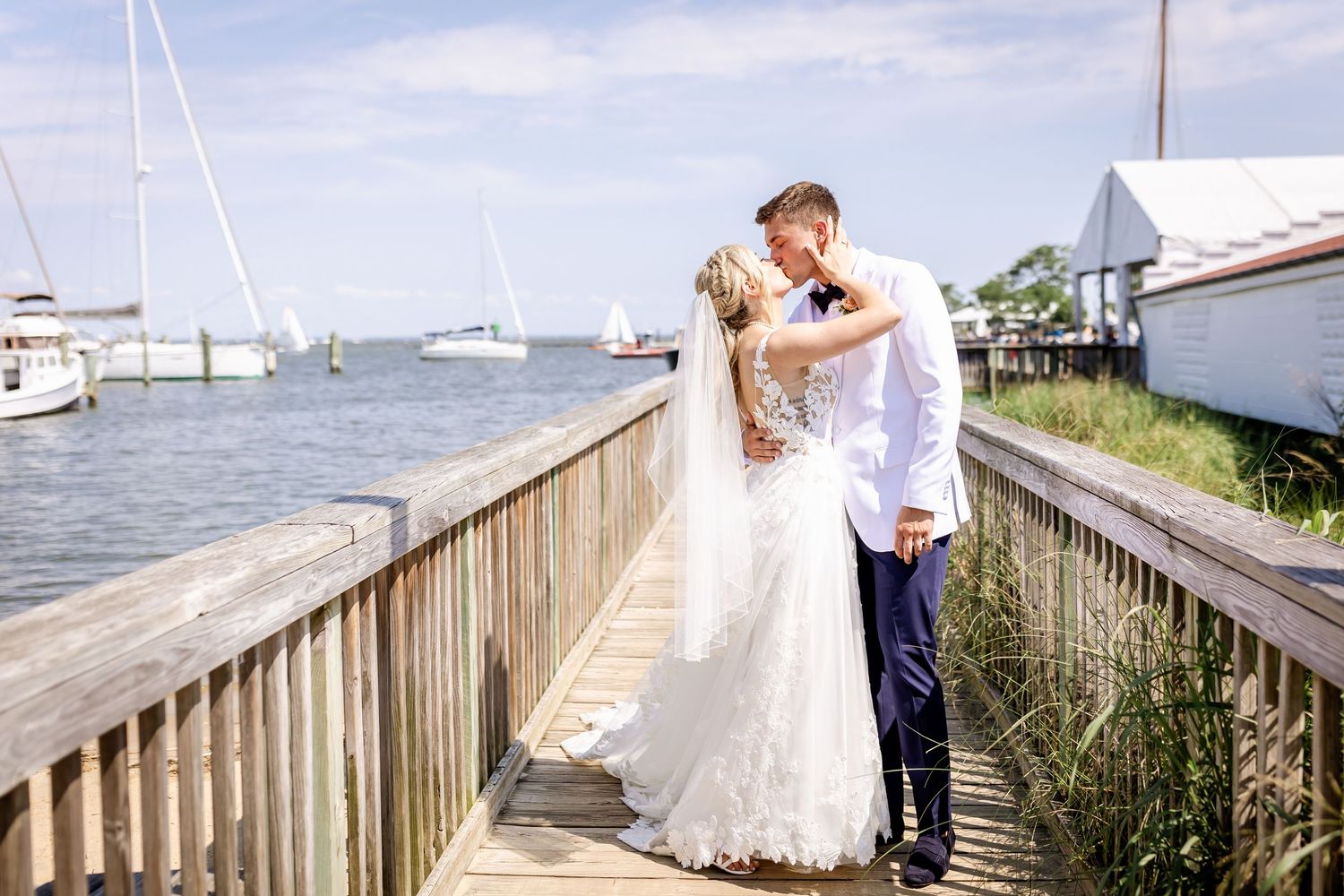 A wedding couple embraces on a marina boardwalk surrounded by boats and water on a sunny summer day.