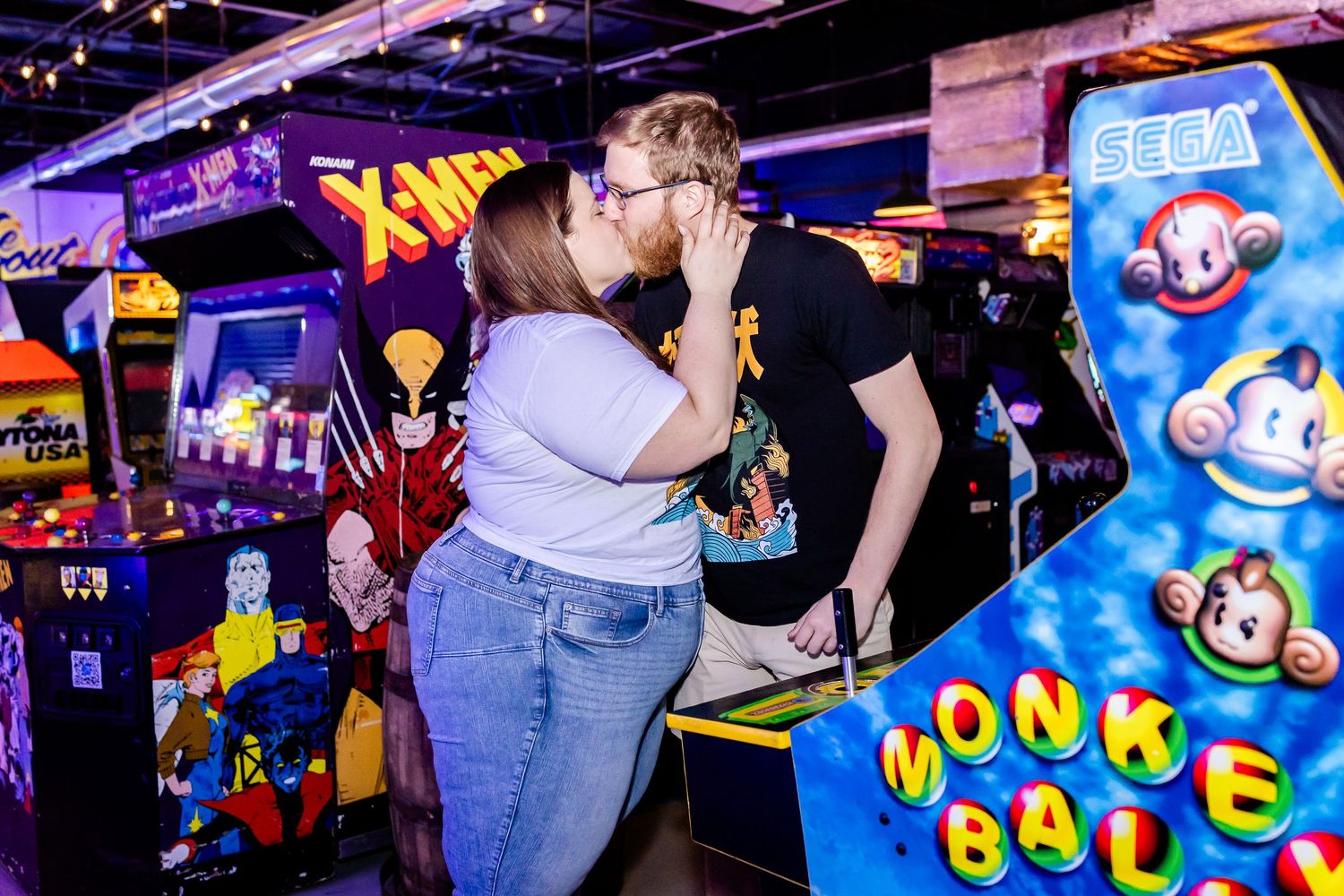 Two people share an intimate moment while playing arcade games at a retro gaming venue with X-Men and Monkey Ball cabinets.