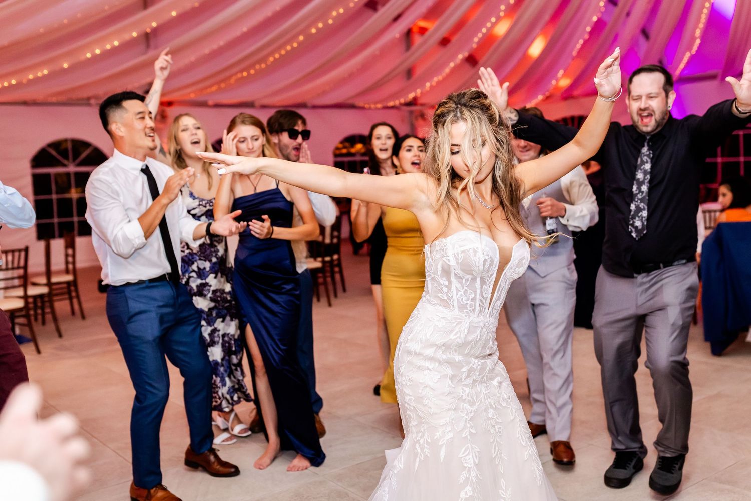 Wedding guests dance and celebrate under a pink-draped tent ceiling at an evening reception celebration.