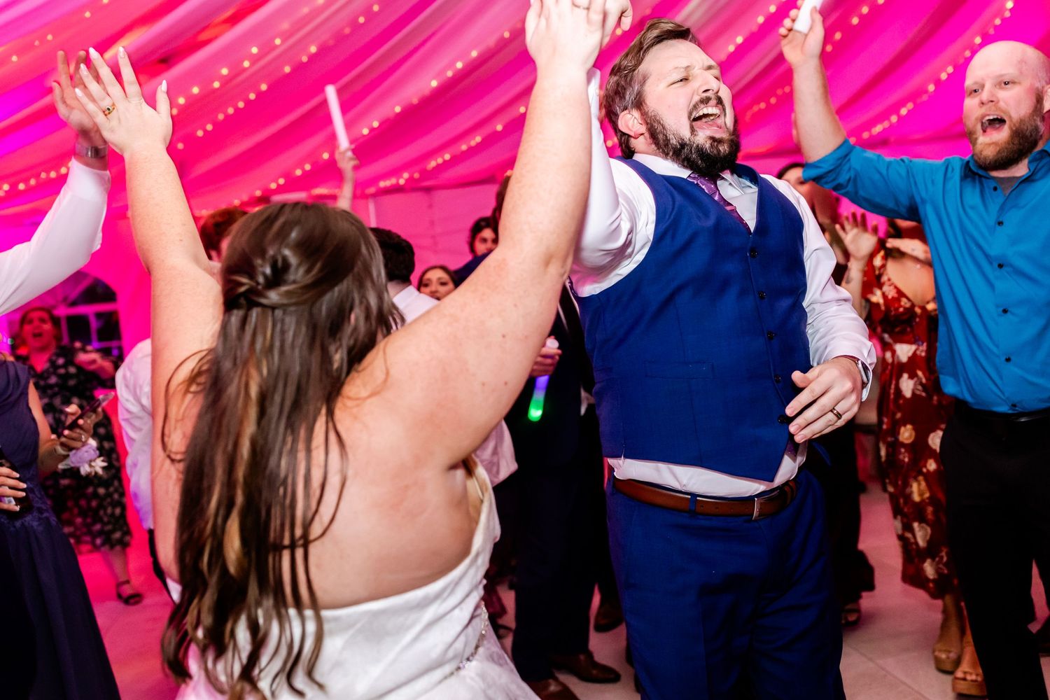 Wedding celebration with energetic dancing and raised arms under pink tent lighting.