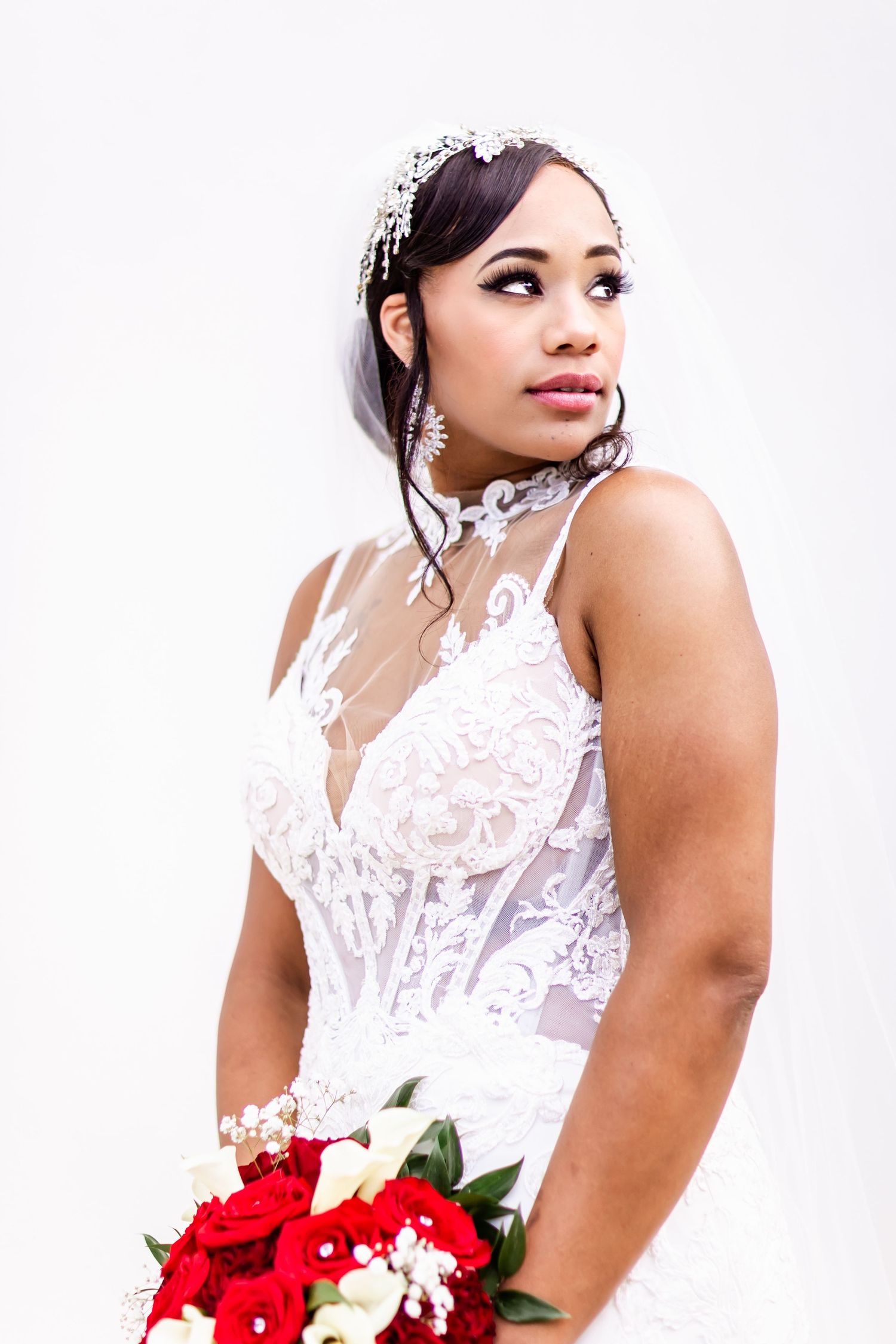 Elegant bride wearing a white lace dress and floral crown holding a red rose bouquet.