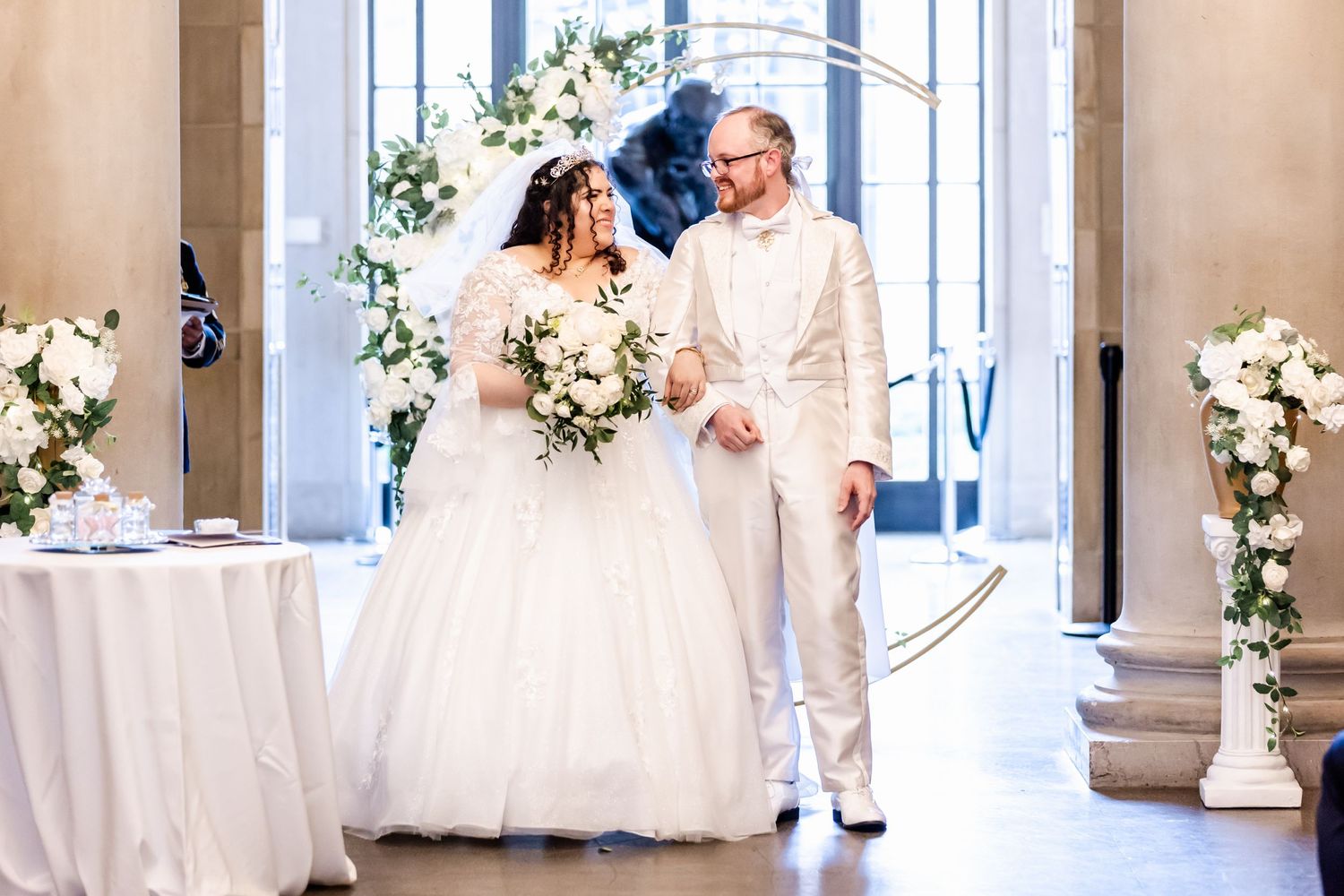 A bride in a white ballgown and companion walking through an elegant venue with floral arrangements.