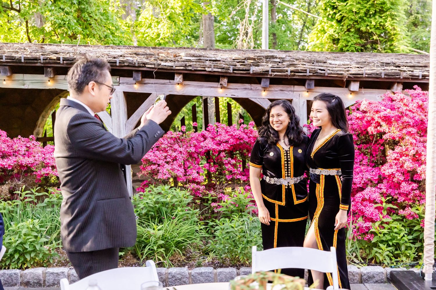 A graduation ceremony takes place in a garden with vibrant pink azalea flowers in the background.