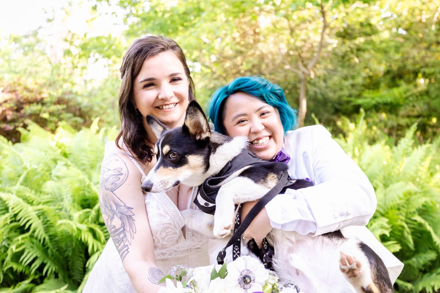 Happy couple holds their black and white dog while surrounded by green ferns in garden setting.