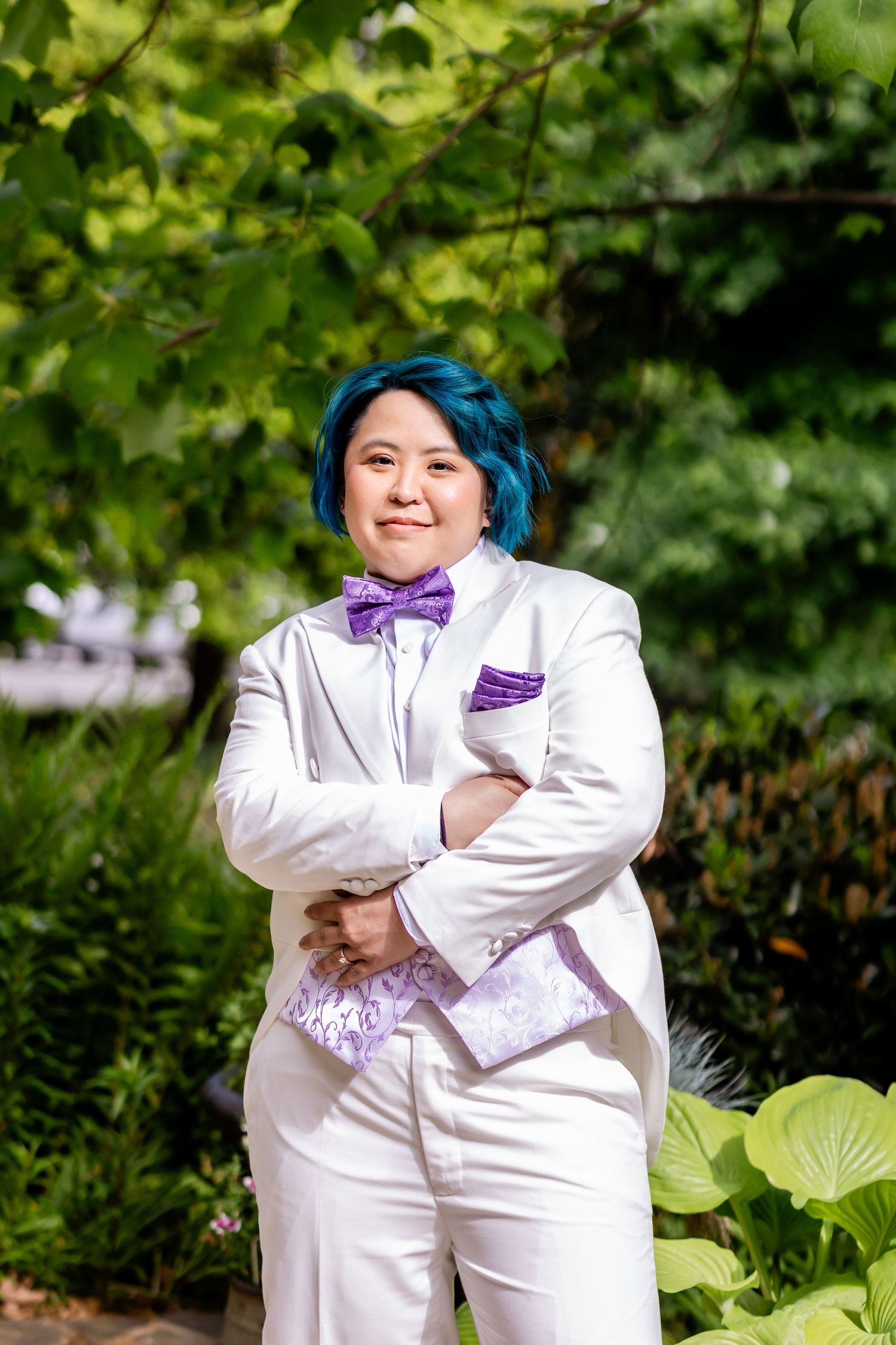 Person in white suit with purple bow tie poses against green garden background.