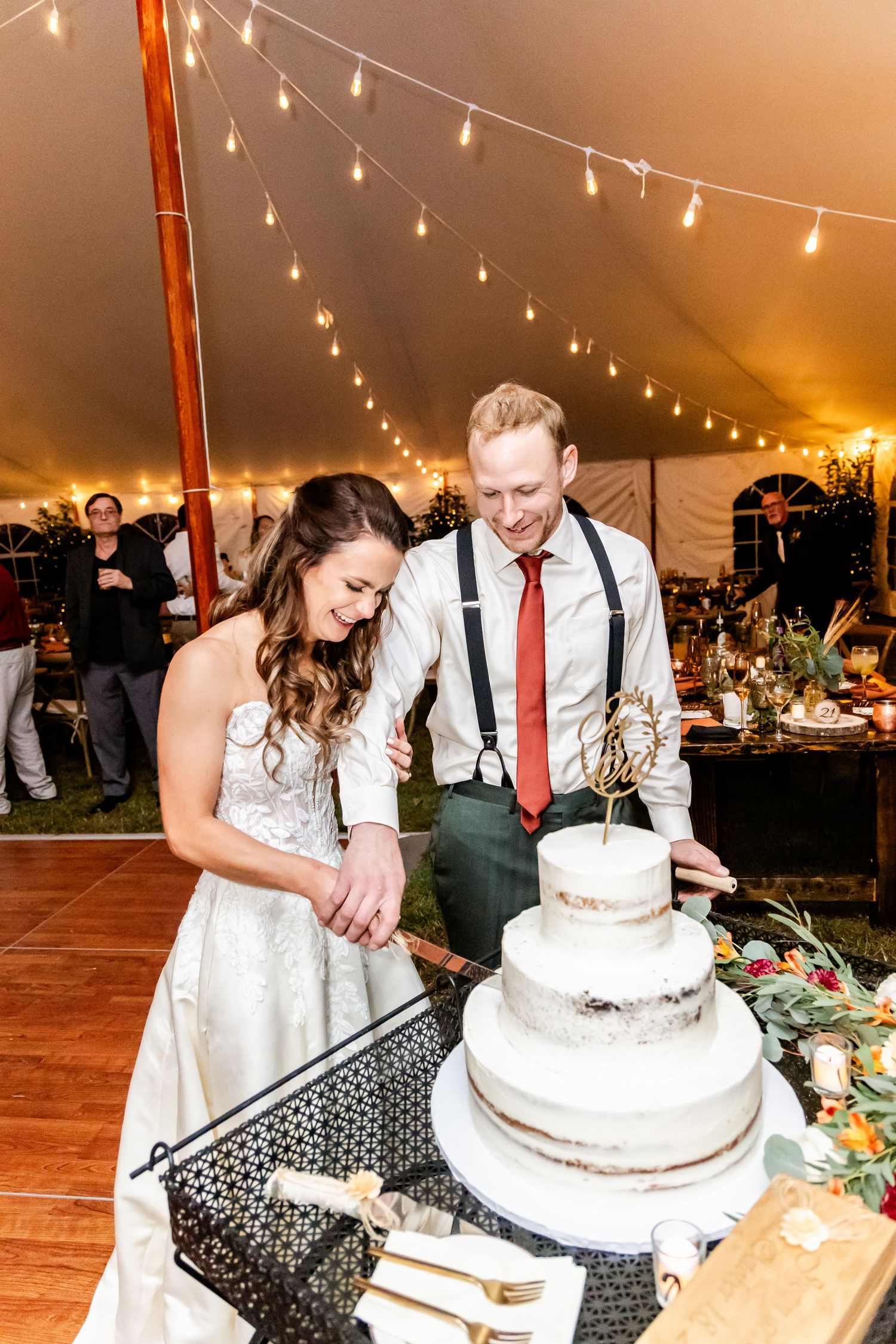 Couple cutting their white wedding cake together under warm string lights in a reception tent.