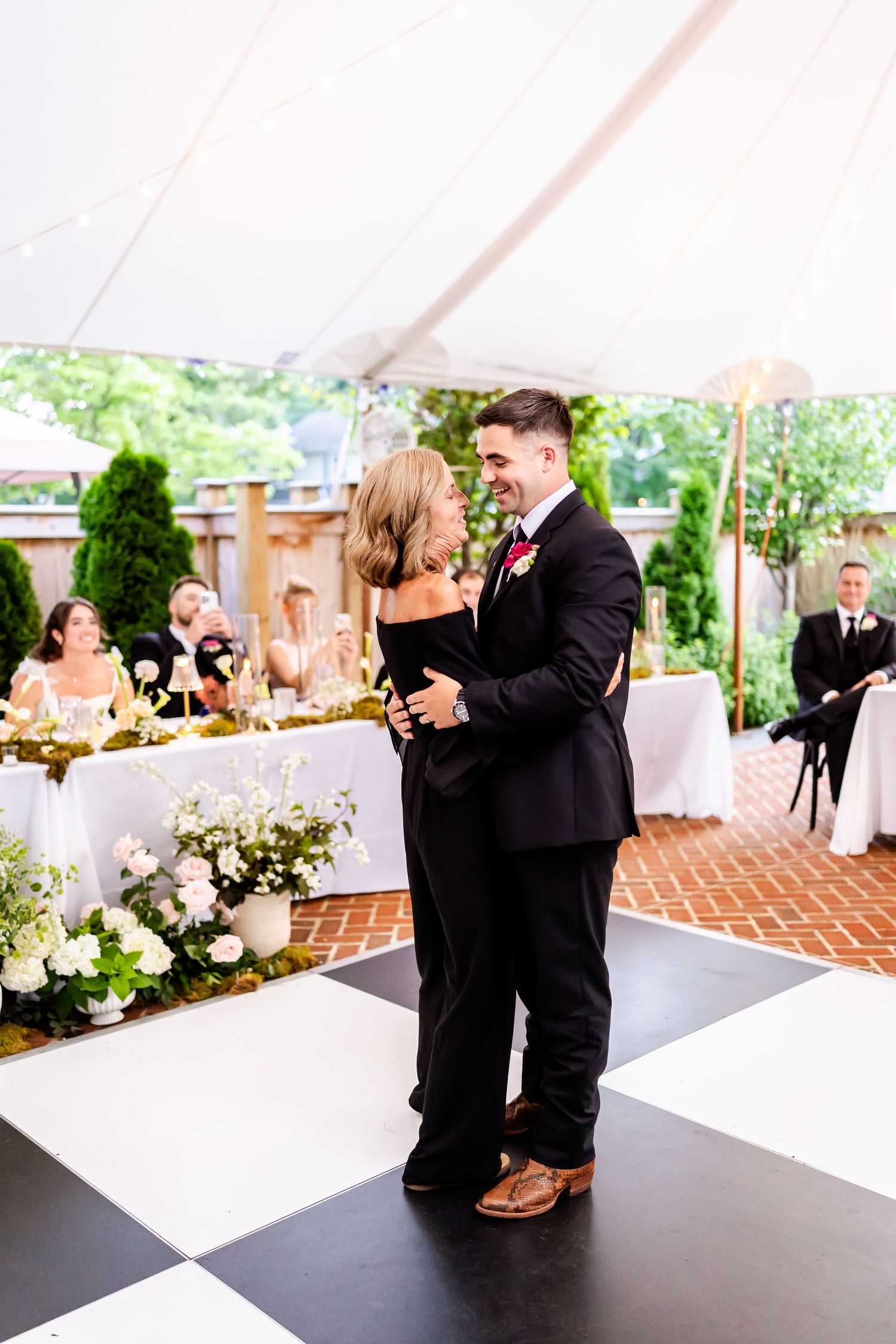 A couple shares their first dance on a black and white checkered dance floor under an elegant outdoor tent reception.