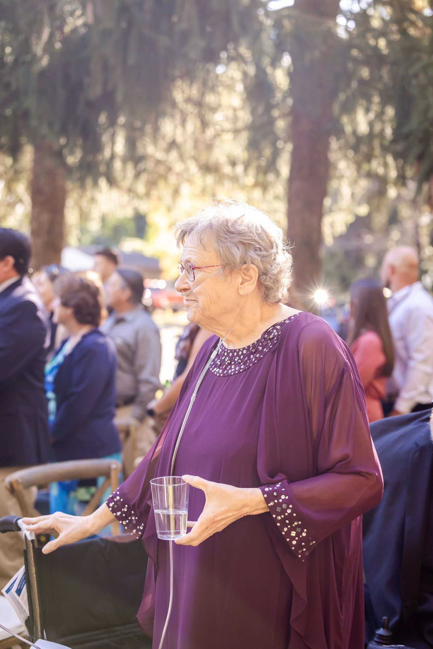 A guest in a purple dress enjoys refreshments during an outdoor event with other attendees visible in the background.