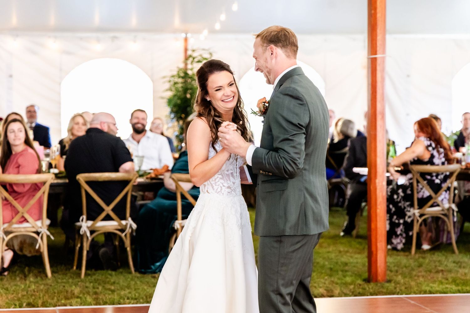 Wedding couple dances surrounded by guests in a tent decorated with romantic string lights.