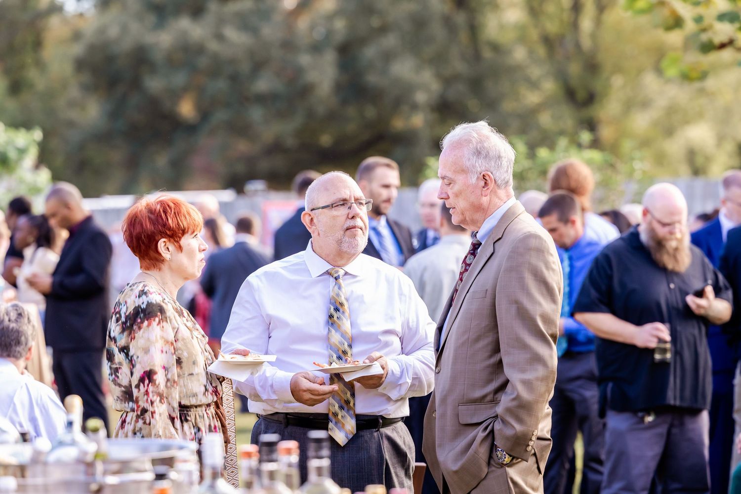 Outdoor wedding reception with guests mingling under trees during daytime celebration.