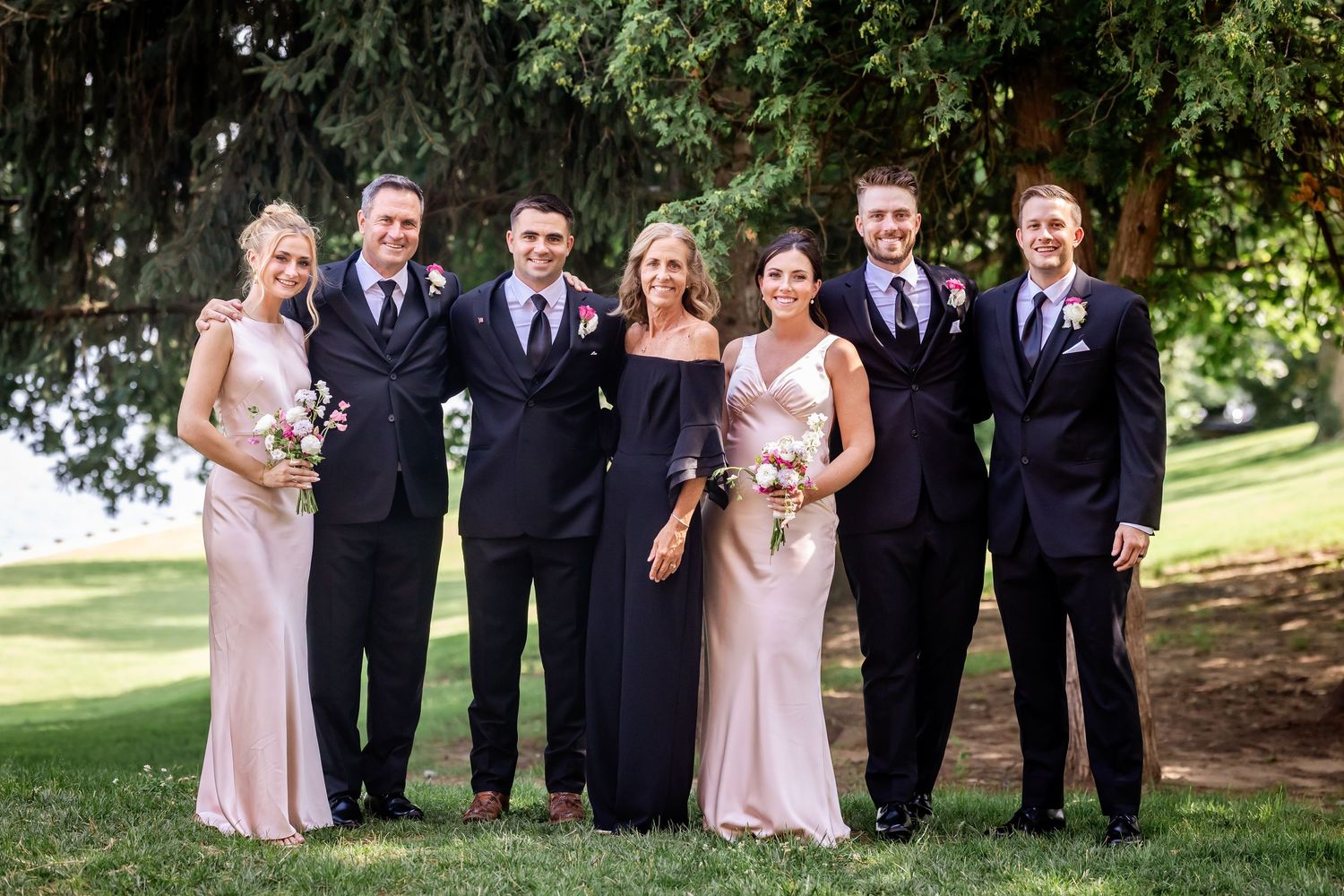 Bridal party in pink dresses and black suits poses together on a grassy lawn.
