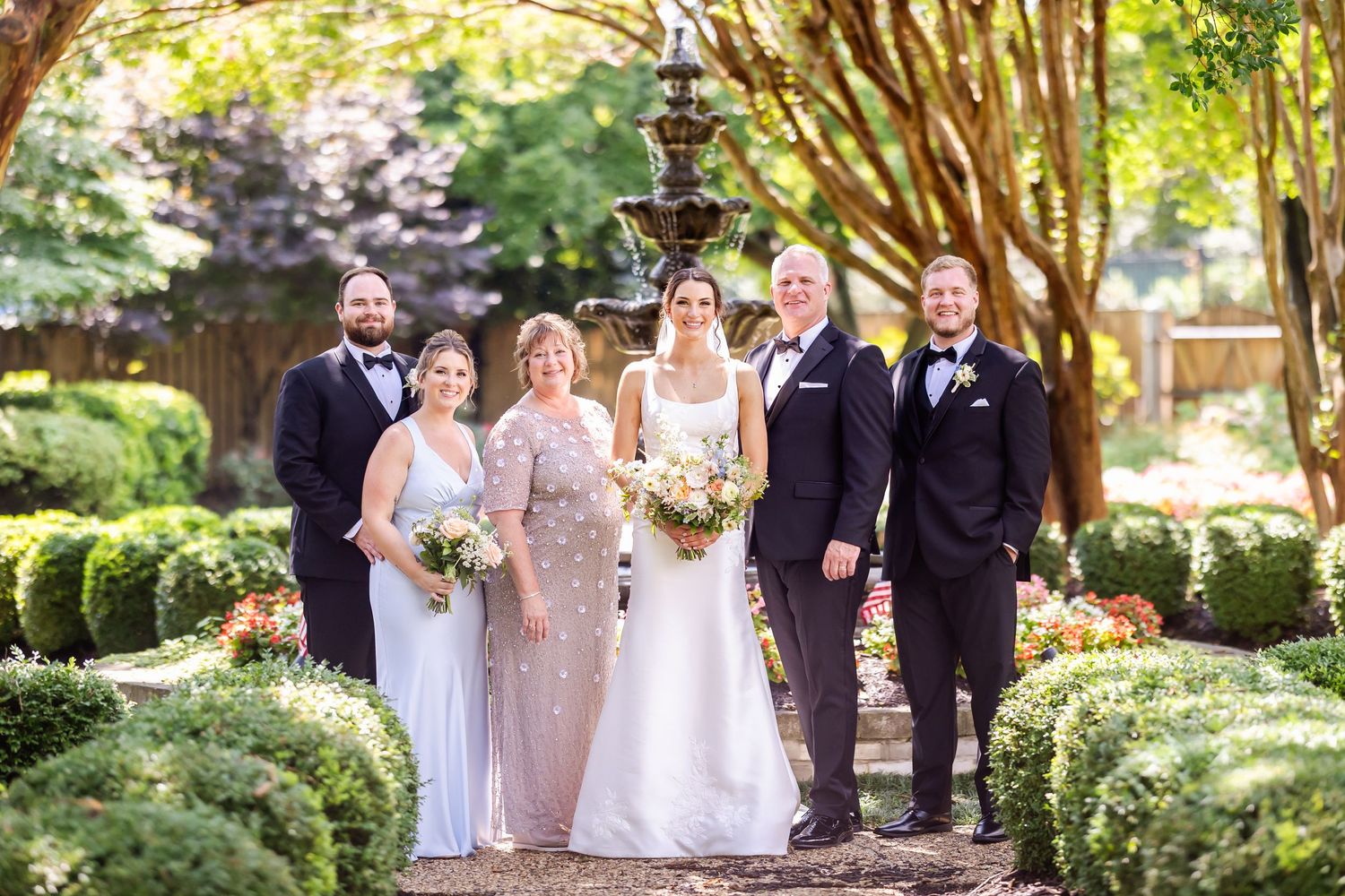 Wedding party poses for formal photo in garden setting with fountain and manicured hedges.