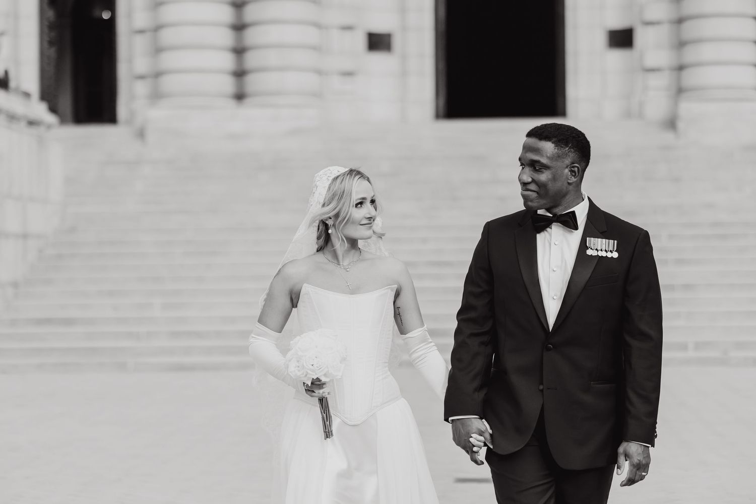 Black and white photo of a bride and groom holding hands walking in front of grand stone steps.