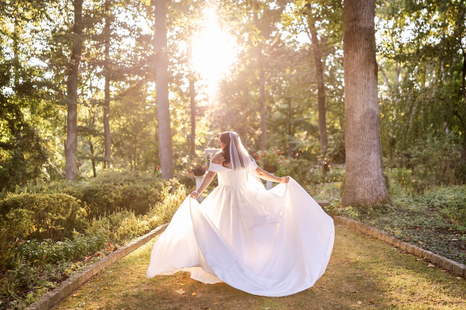 Bride in white gown and veil twirling in sunlit forest garden path on her wedding day.