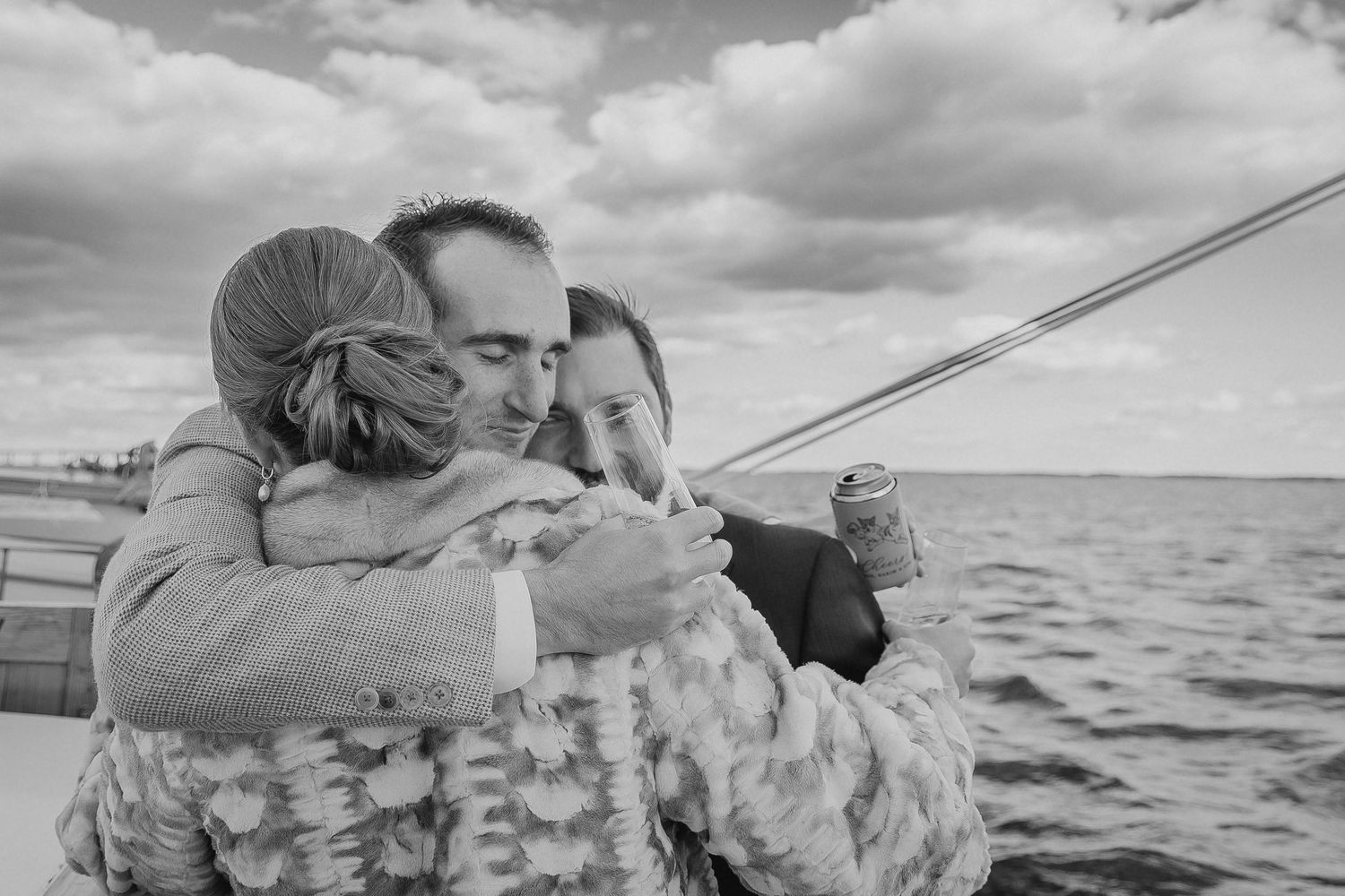 Black and white photo of a couple sharing a warm embrace on a boat with cloudy skies over water.
