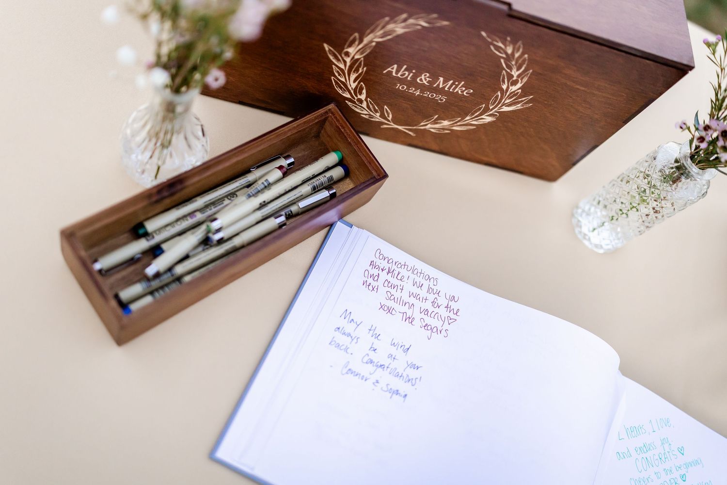Wooden wedding guest book box engraved with Ali & Mike, open with pens, beside a signed open guestbook on a table.