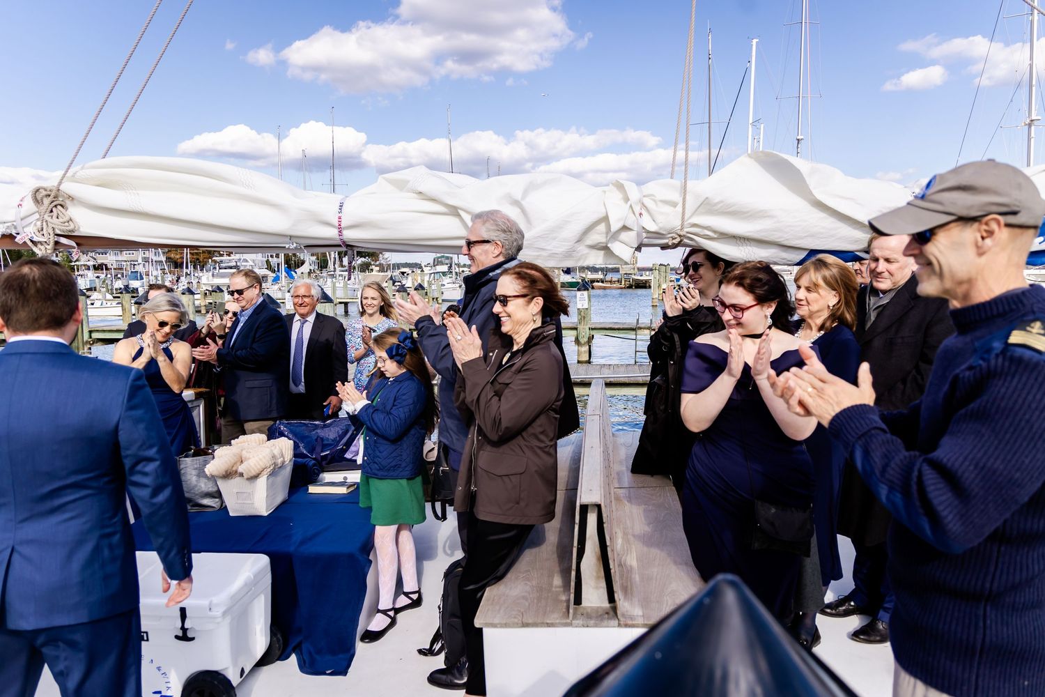 Group of elegantly dressed people applauding on a yacht deck at a marina during a sunny outdoor ceremony.