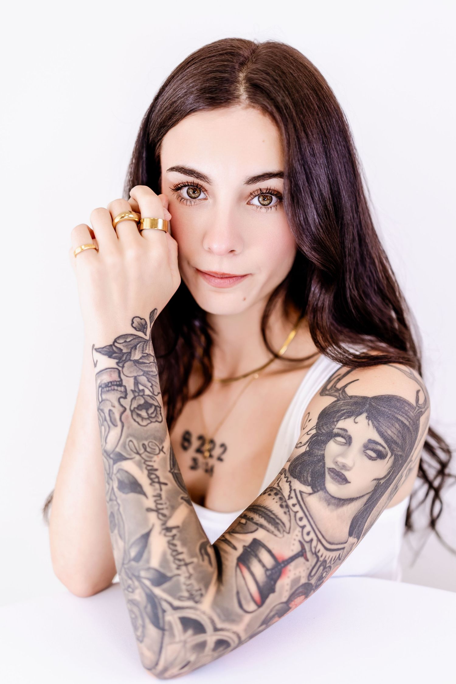 Young woman with long dark hair, gold rings, and full sleeve tattoos poses against a white background.