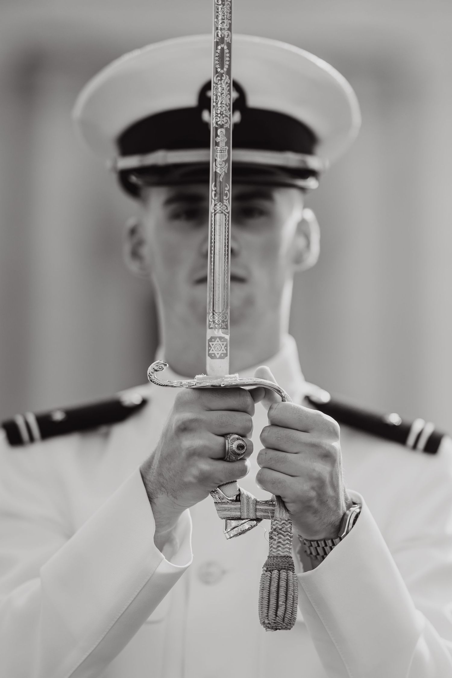 Naval officer in white dress uniform holding a ceremonial sword vertically in a black and white photo.