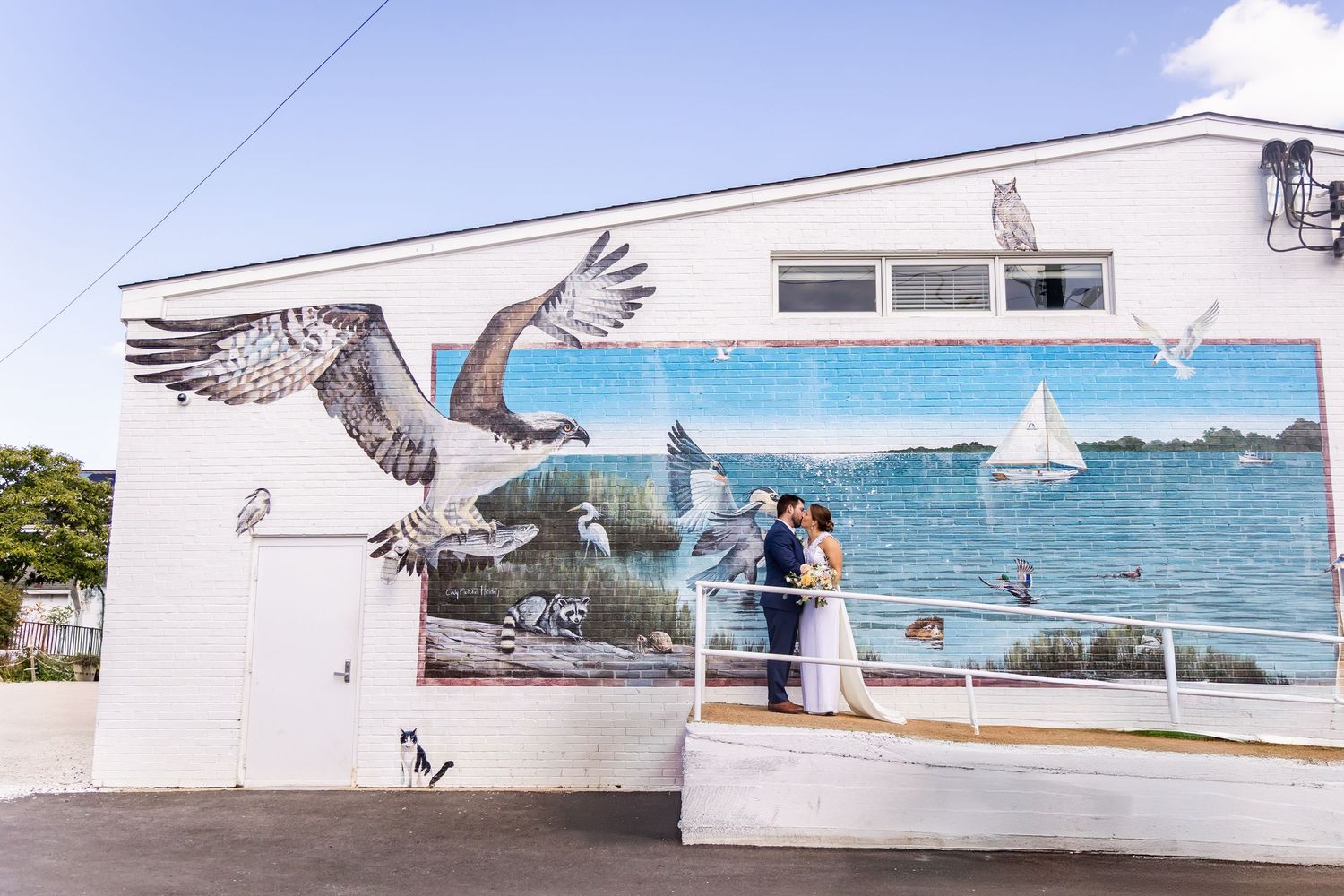 Couple embracing in front of a colorful coastal mural featuring ospreys and sailboats on a white building.