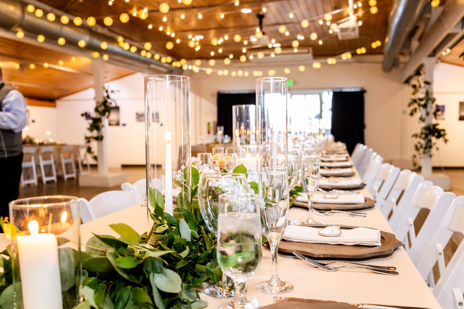 Elegant wedding reception table with greenery runner, candles, glassware, and string lights overhead in a bright venue.