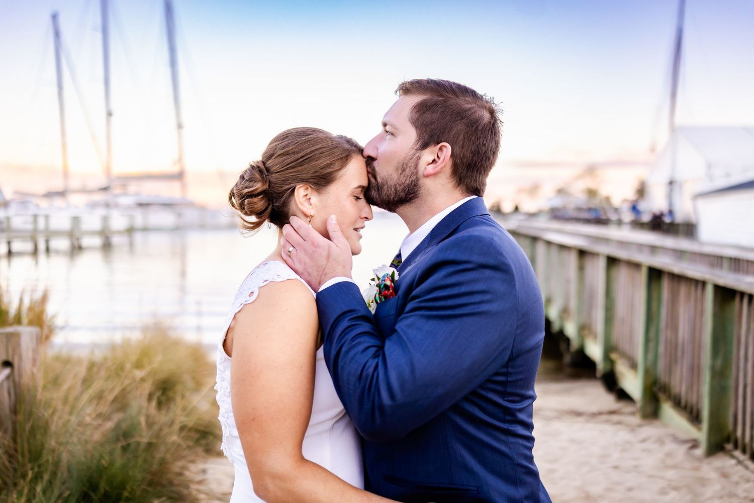 A groom in a navy suit tenderly kisses his bride in a white lace dress near a waterfront dock at sunset.
