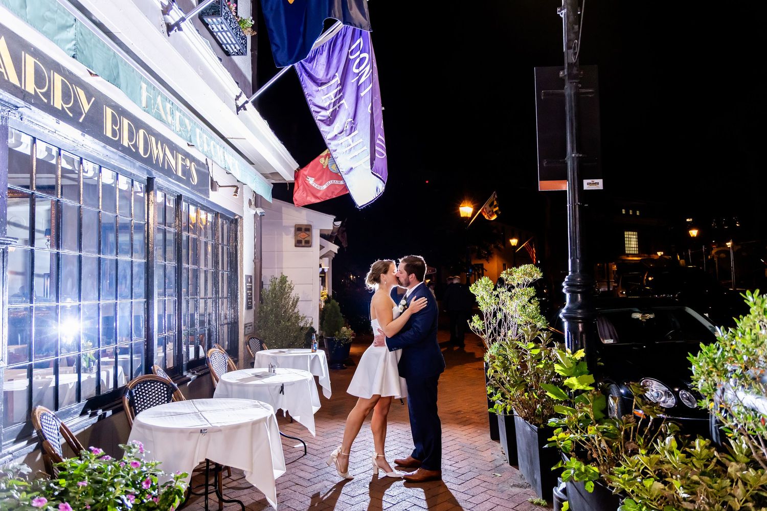 Couple sharing a romantic kiss outside a French restaurant at night, surrounded by bistro tables and string lights.