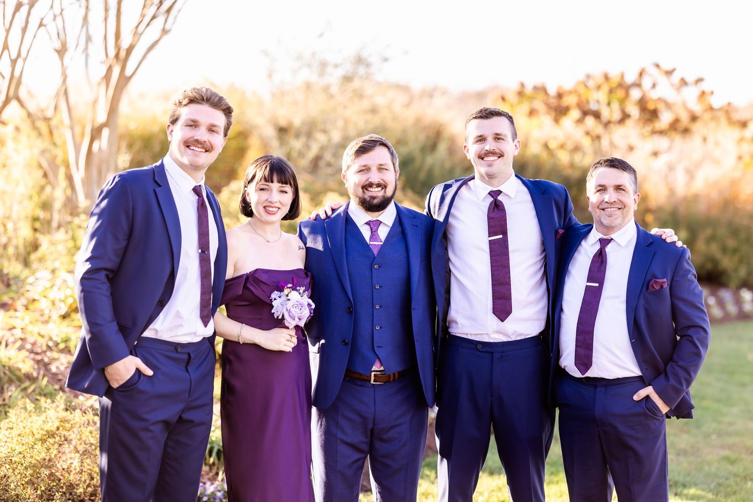 Wedding party in navy suits and purple dress pose together outdoors during sunset.