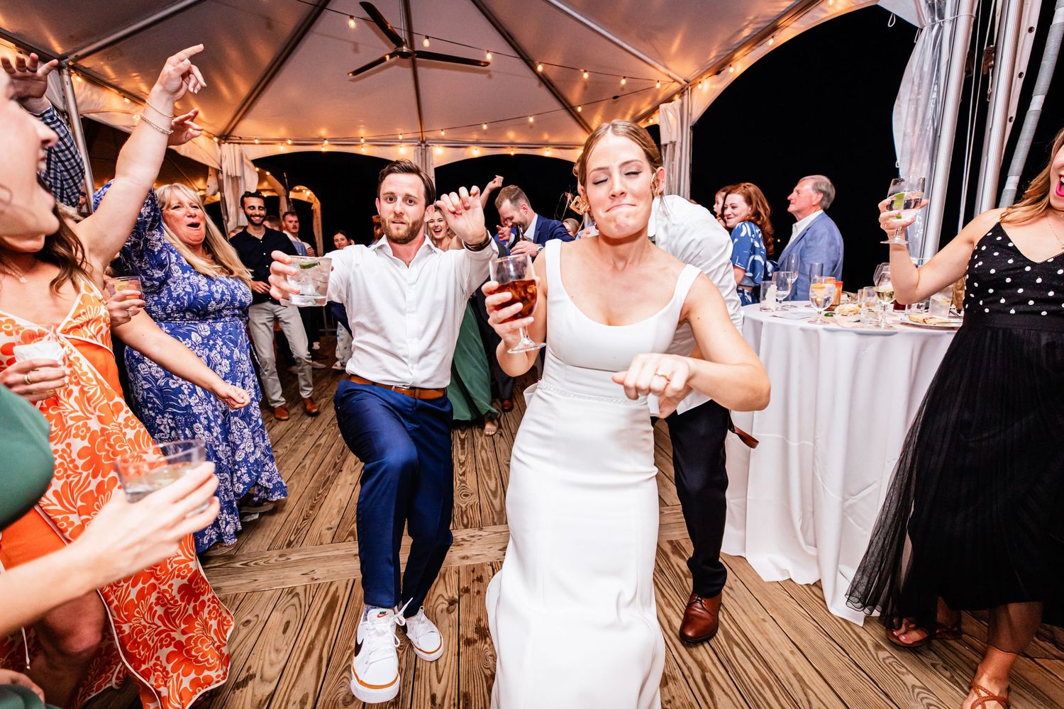 Wedding reception party guests dancing and celebrating with the newlyweds under a tent canopy at night.