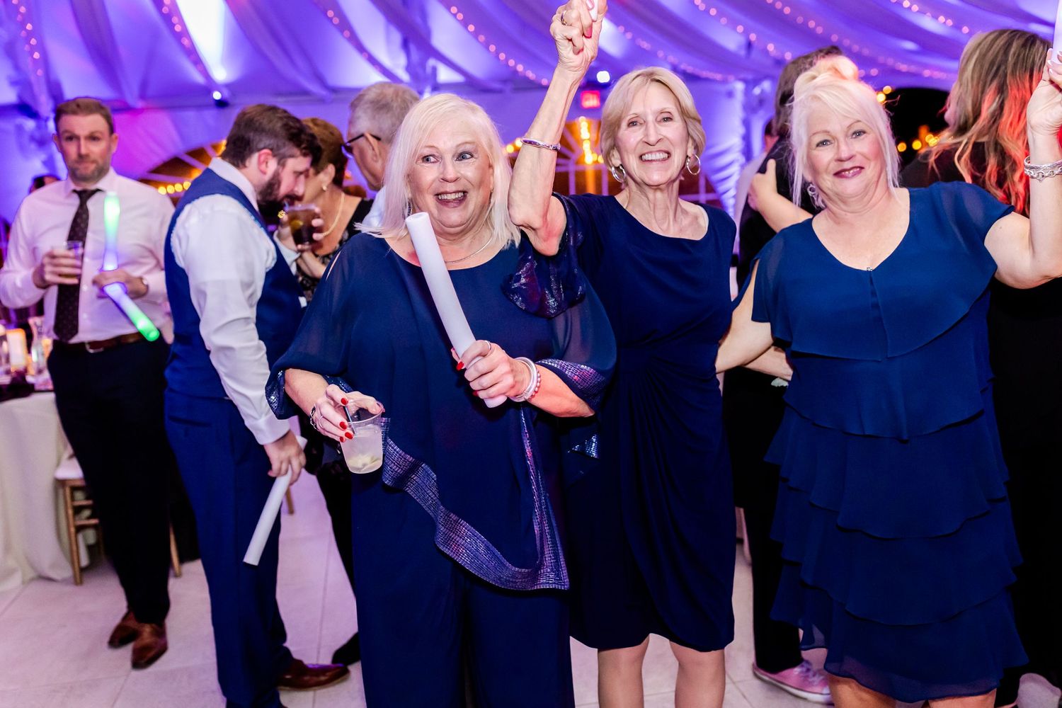 Wedding guests in navy blue dresses dance joyfully under purple lighting at an elegant reception.