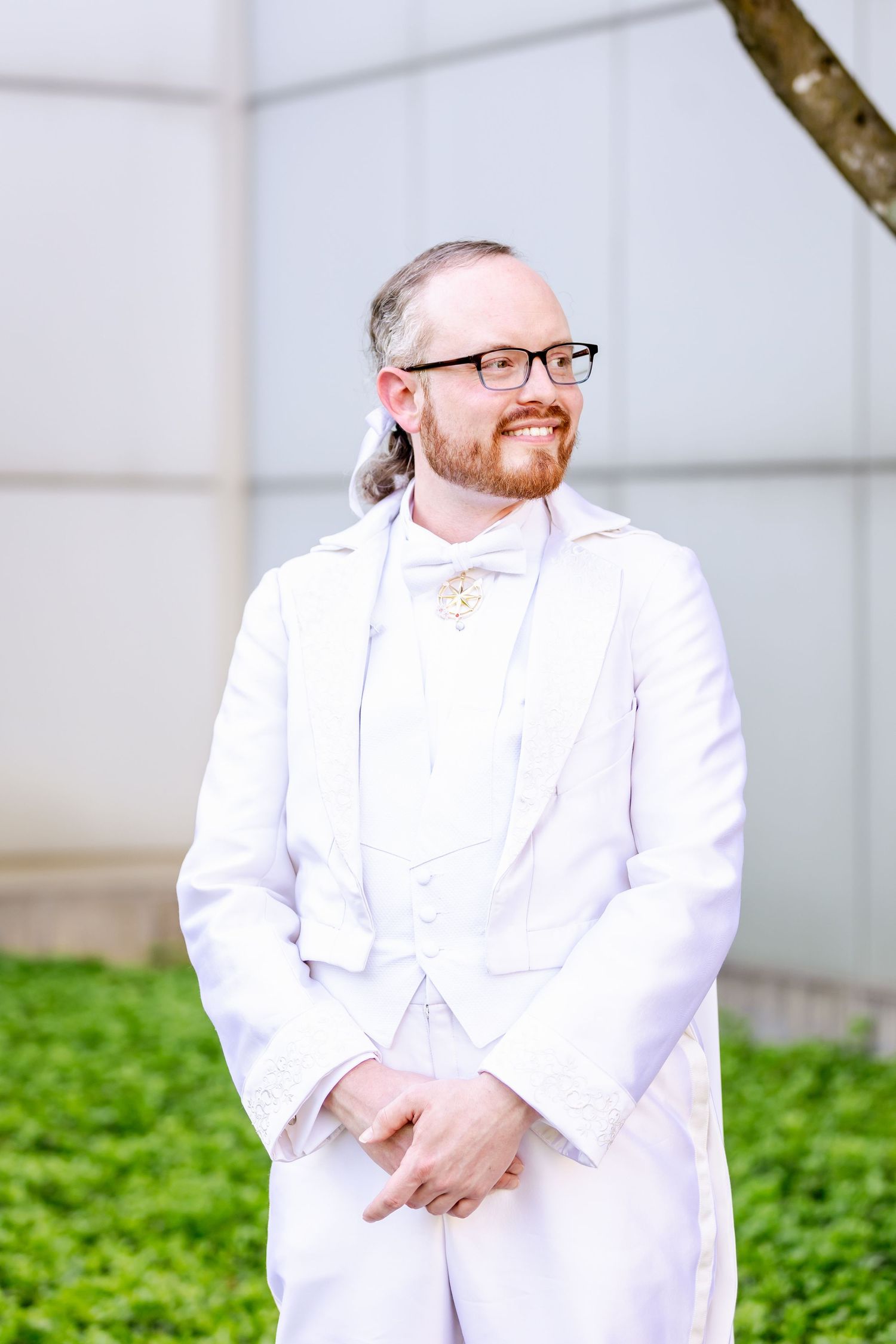 Person in all-white formal wear and glasses stands against white wall with greenery.