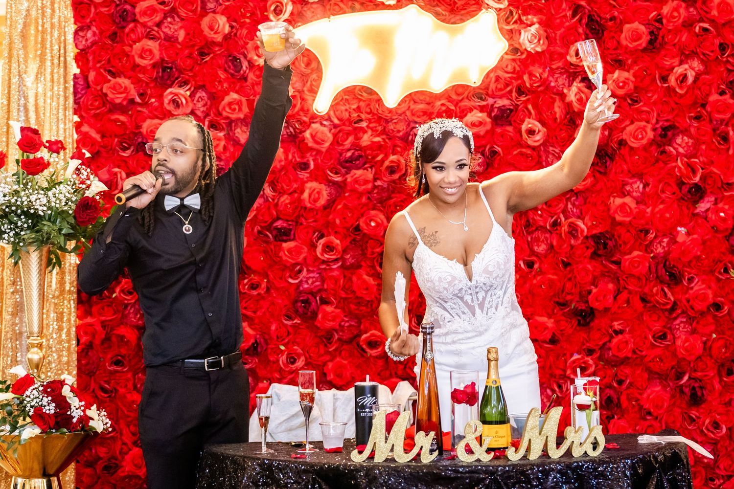 Newlyweds celebrate with champagne in front of a stunning red rose wall backdrop at their wedding reception.
