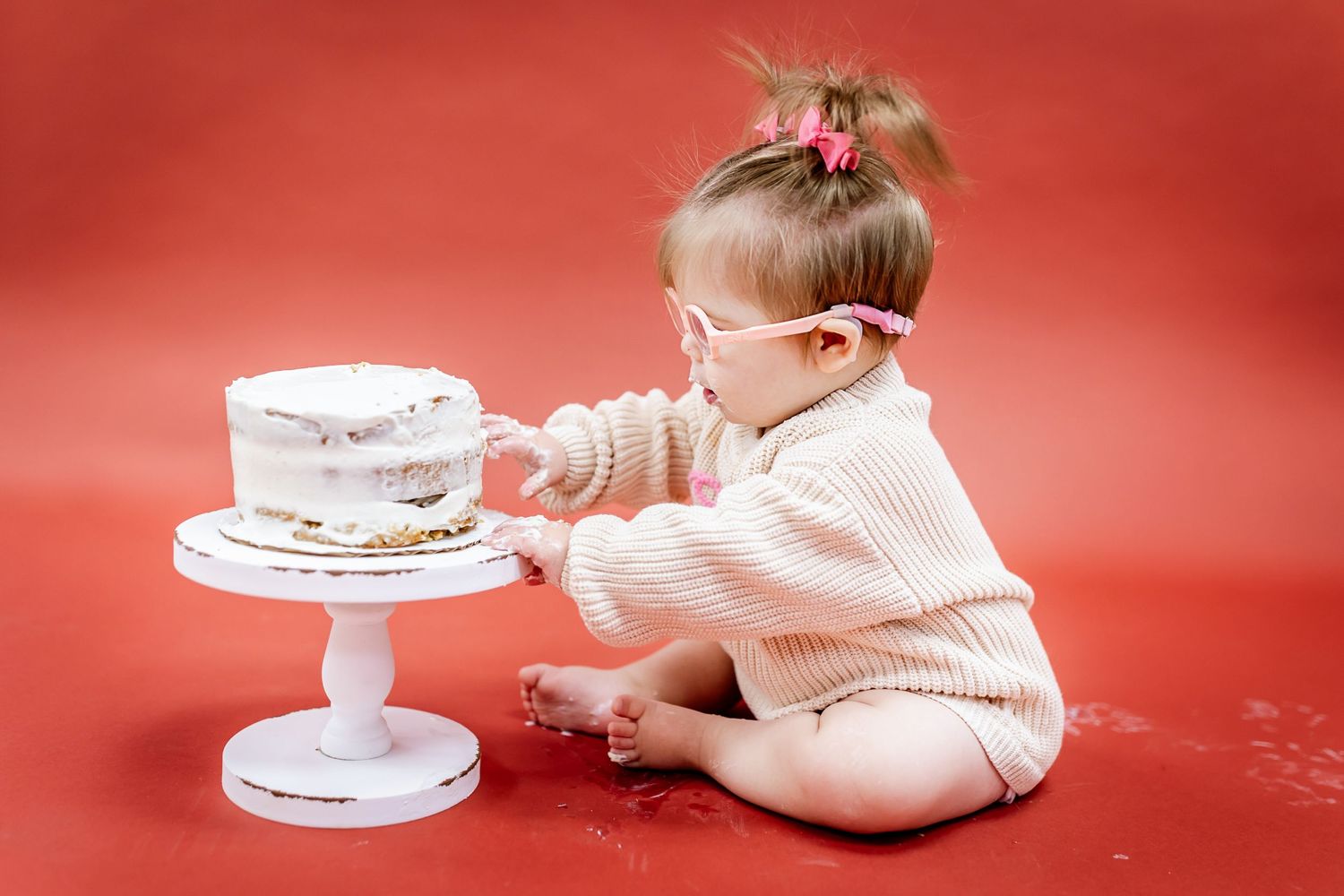 Baby examines white cake on pedestal while sitting on red background.