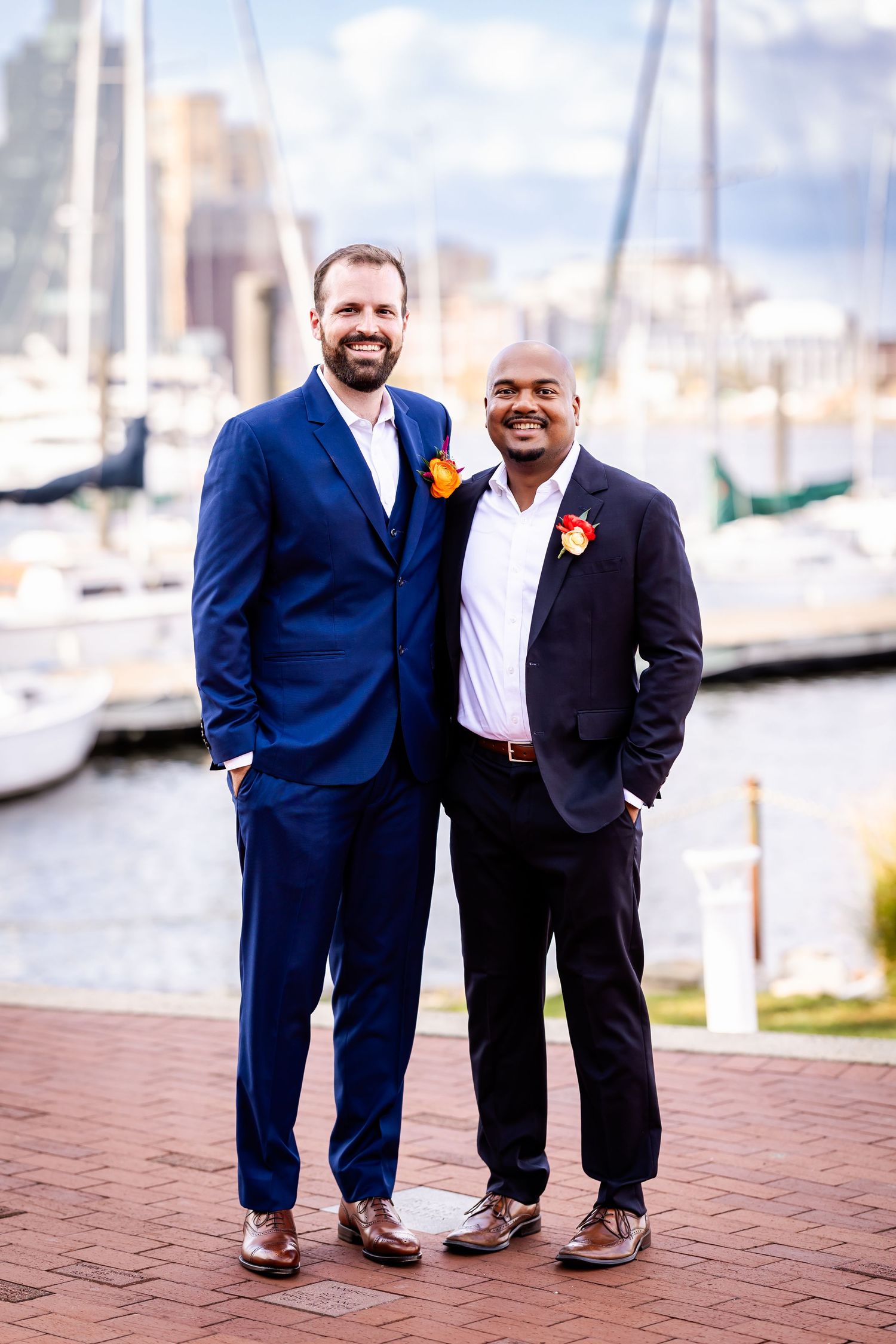 Two stylishly dressed men pose together on a marina boardwalk with boats in the background.
