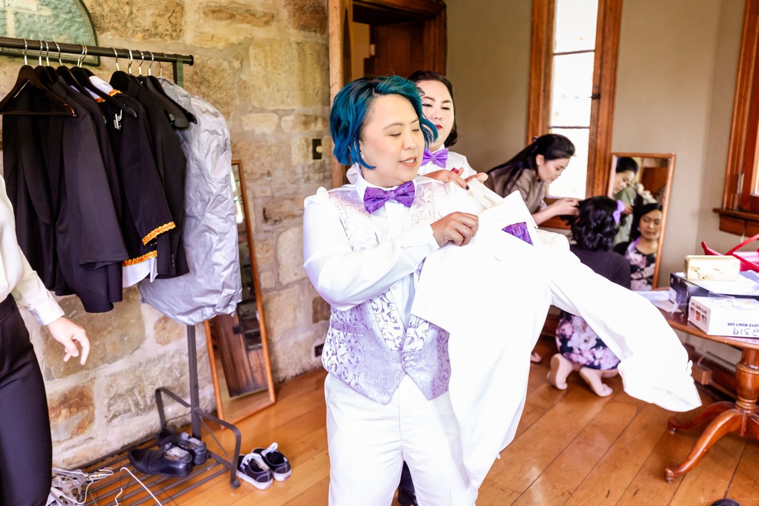 A person in a white suit and blue hair poses playfully during wedding preparations.