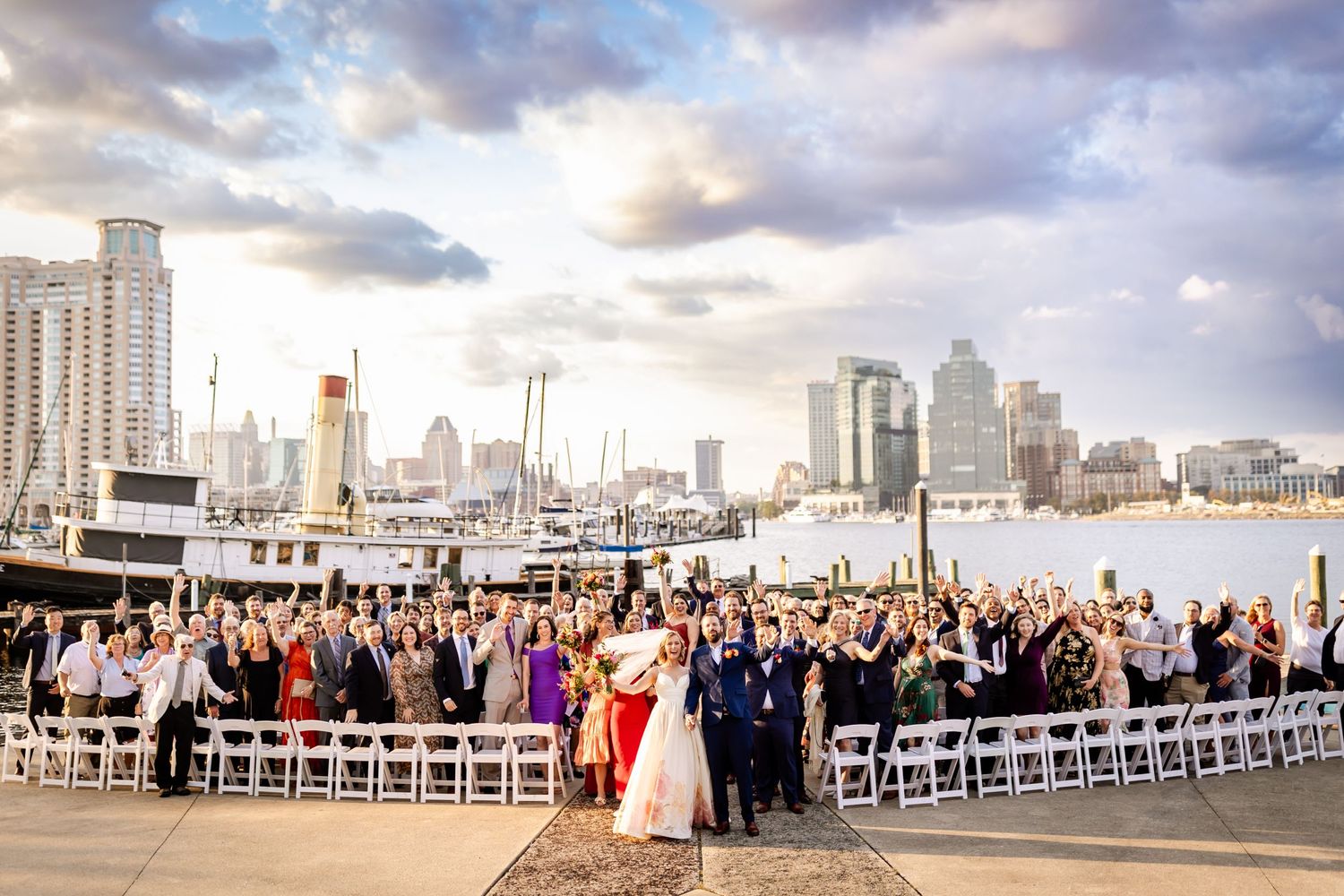 Waterfront wedding ceremony with city skyline backdrop and crowd of seated guests at sunset.
