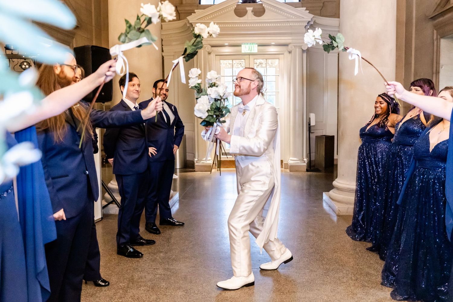 Wedding party creates a joyful tunnel as couple walks through an elegant hallway with columns.
