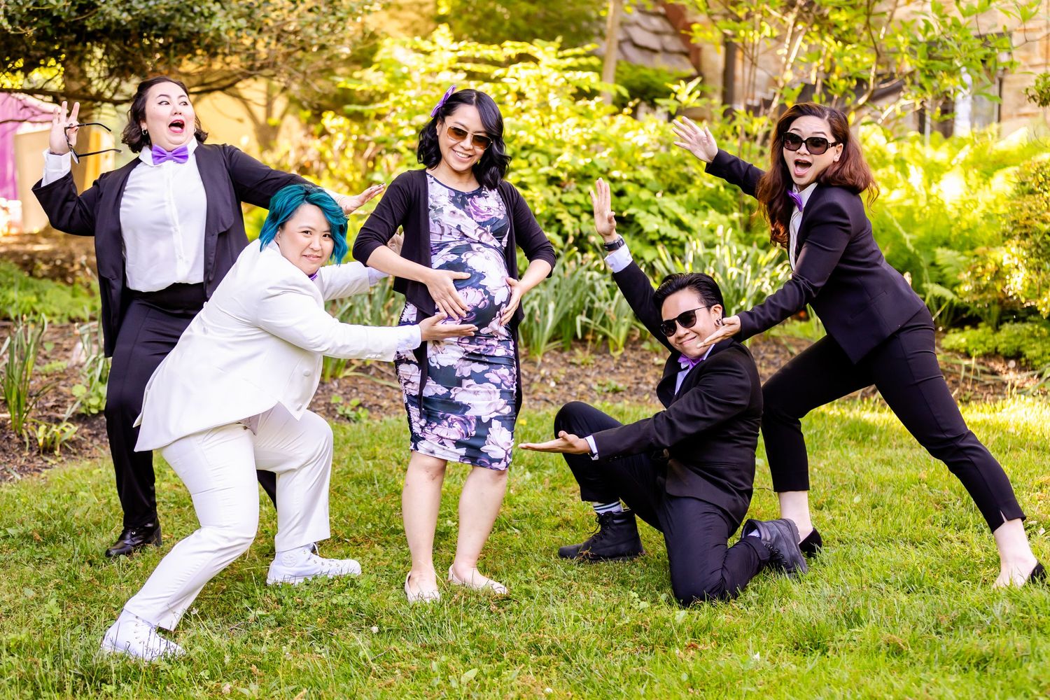 Wedding party striking dramatic action poses in formal attire on a grassy lawn with sunlit trees.