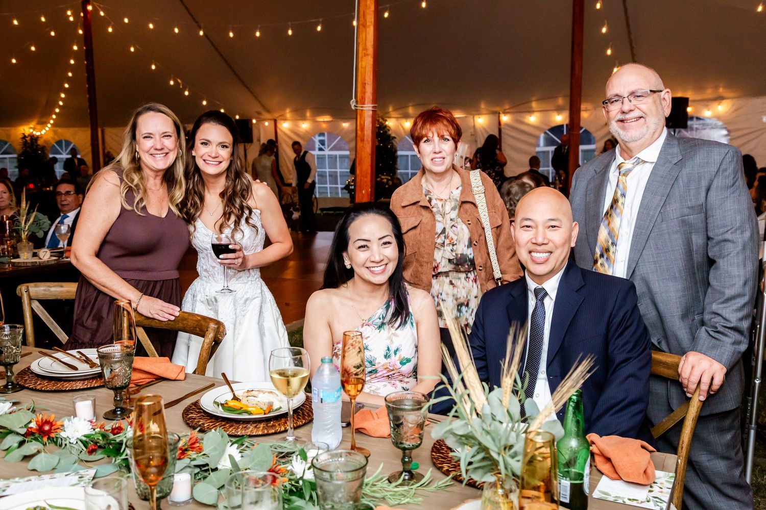 Wedding reception guests enjoying dinner and drinks under string lights in an elegant tent setting.