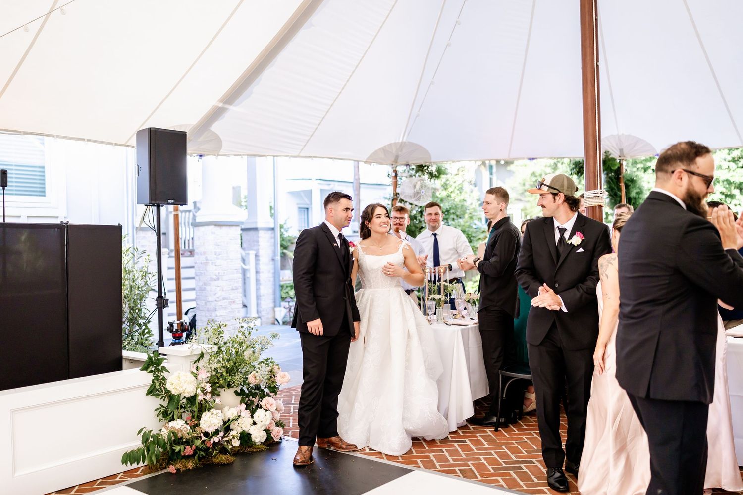 Wedding guests mingle and dance under a white tent at an outdoor venue.