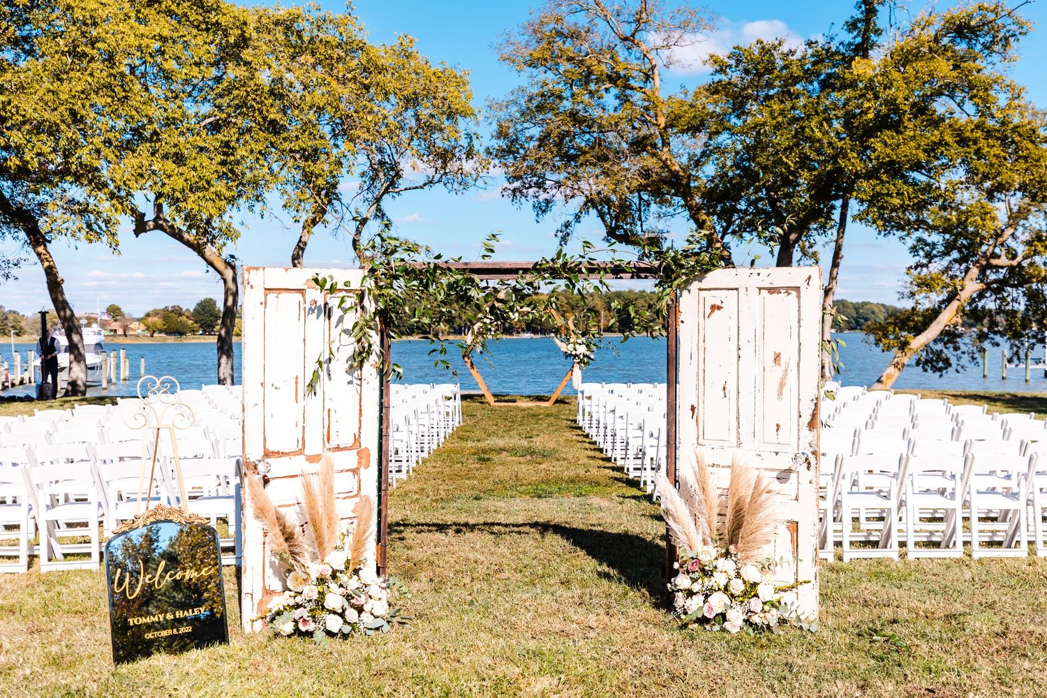 White chairs and floral arrangements line a scenic outdoor wedding ceremony setup overlooking a waterfront view.