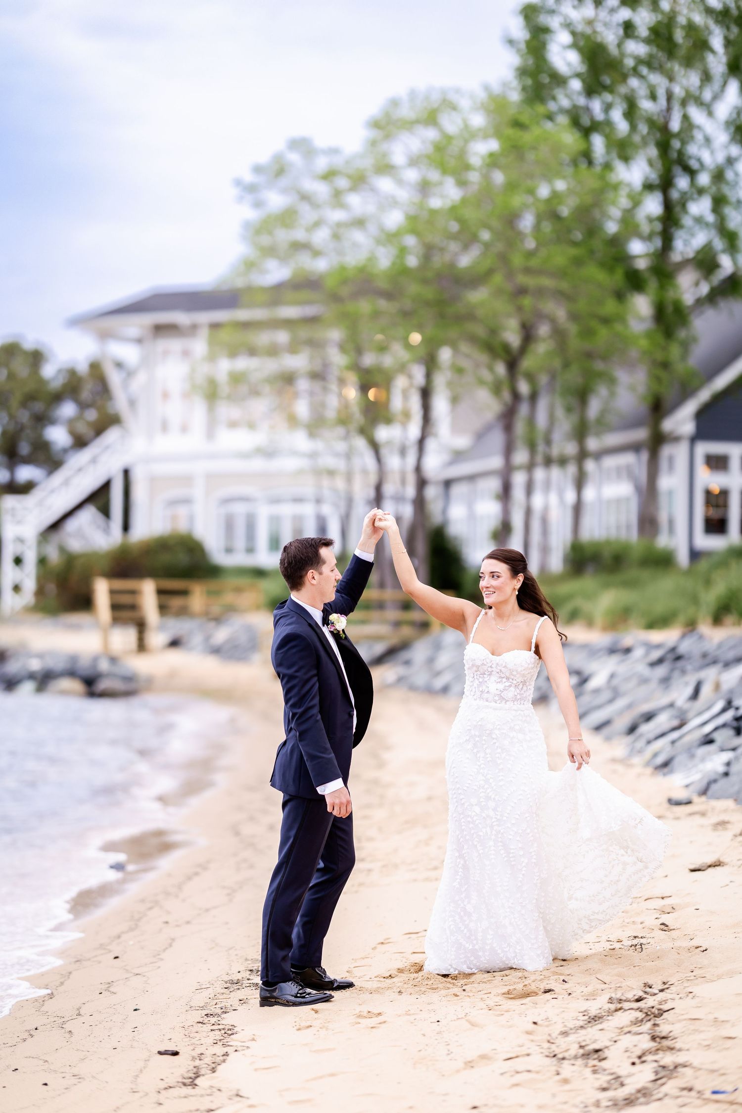 A couple dancing and twirling on a sandy beach with a white coastal home in the background.
