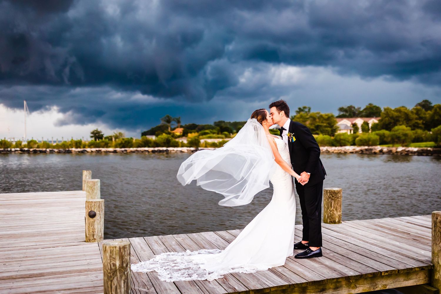 Dramatic waterfront wedding photo on wooden dock with flowing white veil against stormy dark clouds over lake.