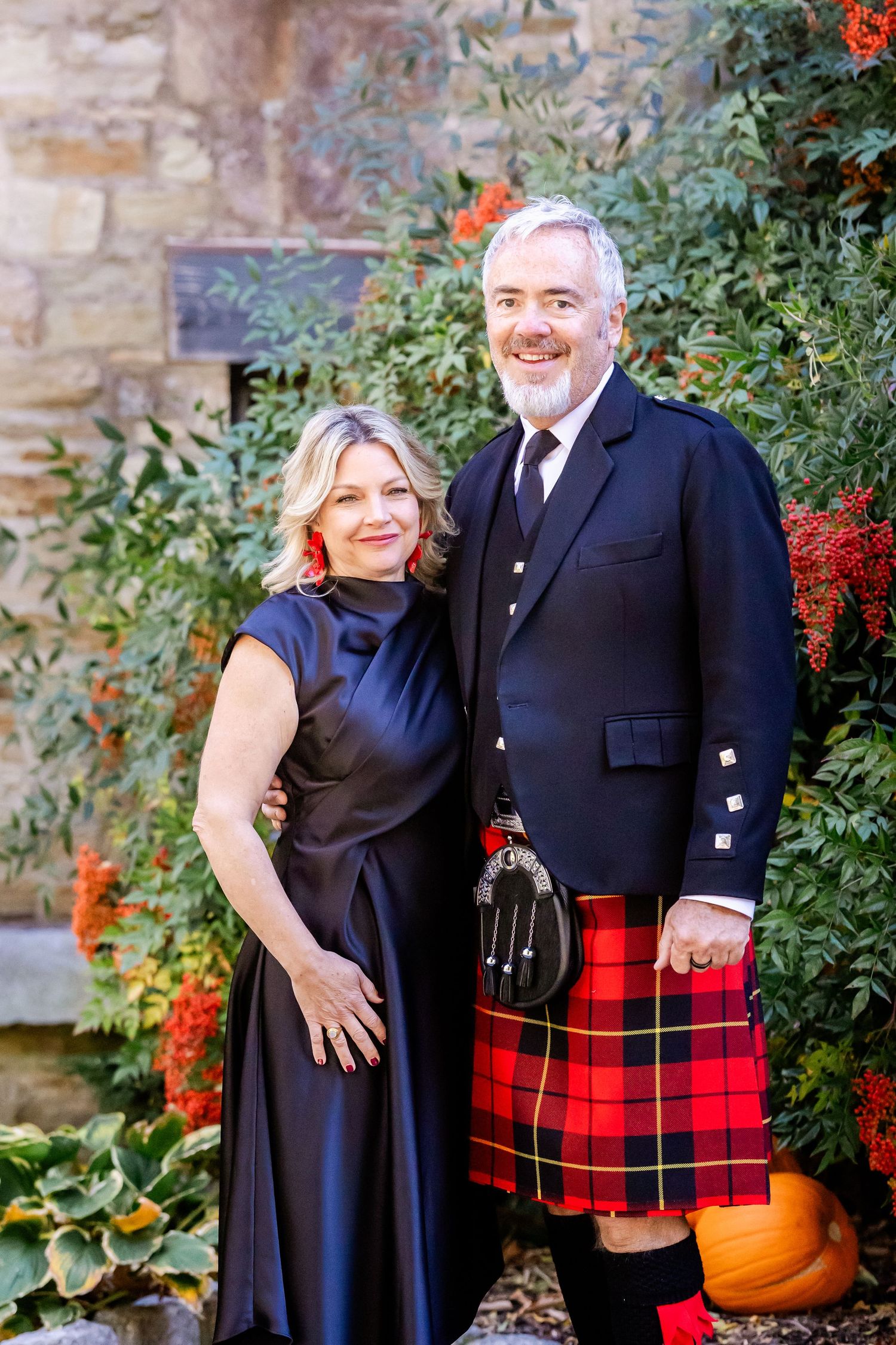 Couple poses outdoors, man wearing traditional Scottish kilt and woman in navy dress.