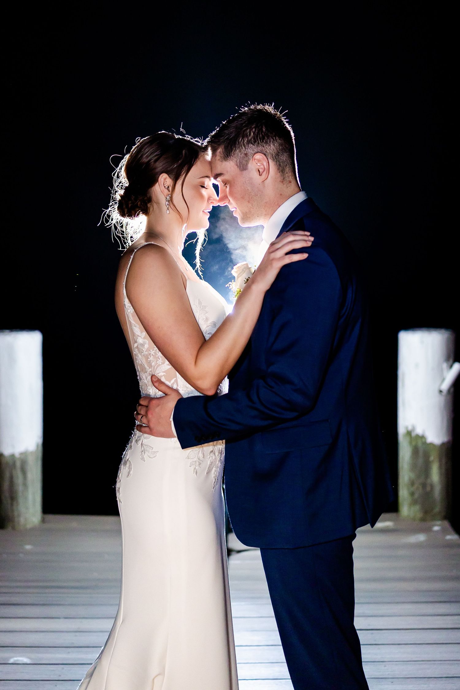 Romantic nighttime portrait of a couple in wedding attire embracing on a dock with dramatic backlighting.