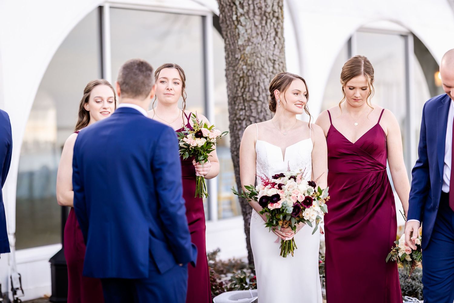 Wedding party members in burgundy dresses and blue suits pose together with floral bouquets.
