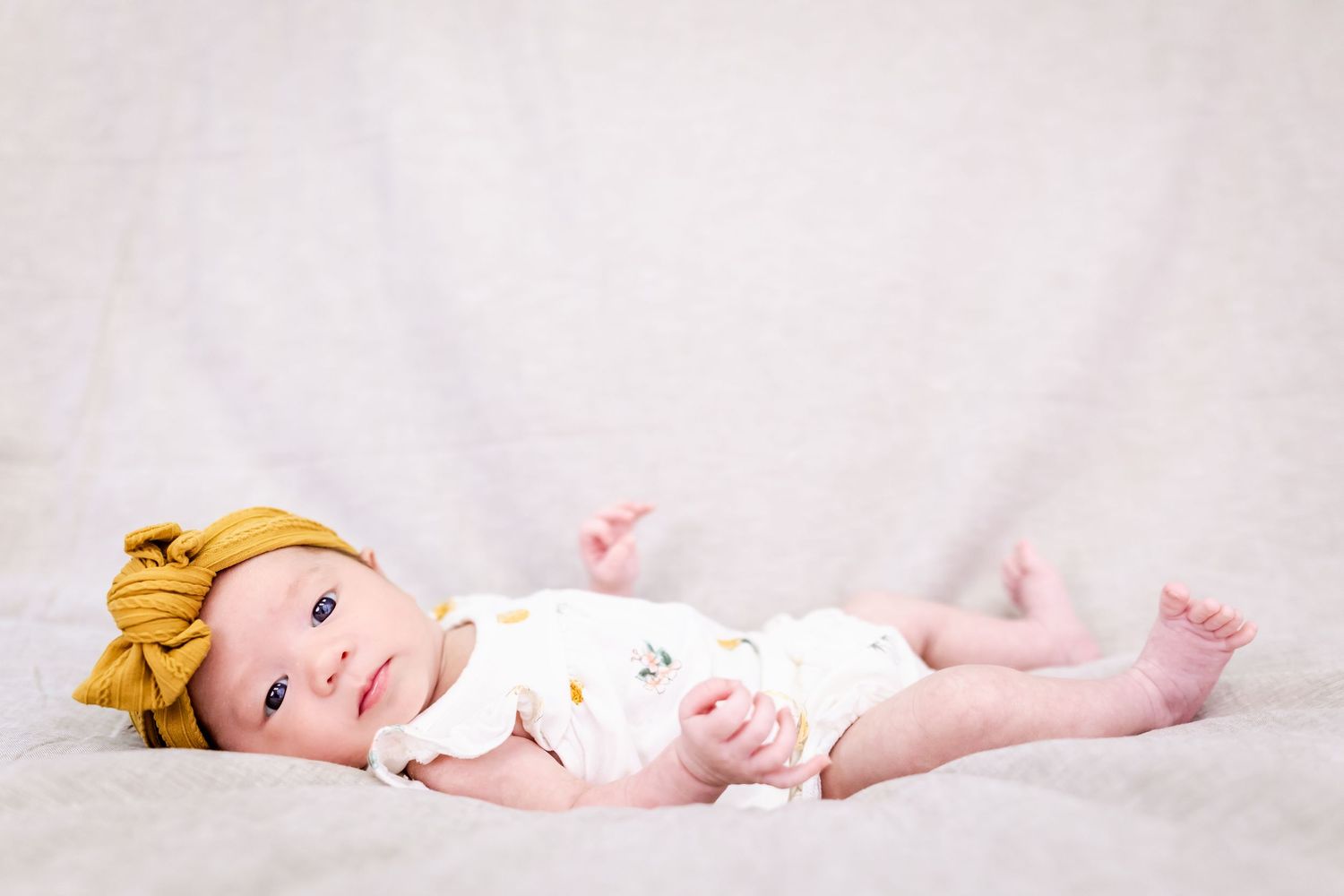 A baby wearing a yellow bow headband lies on soft white fabric.