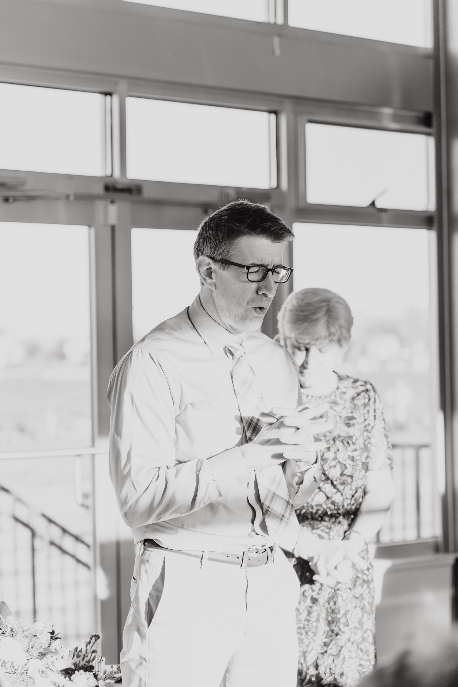 A black and white photograph of a couple in formal attire standing by large windows in an indoor setting.