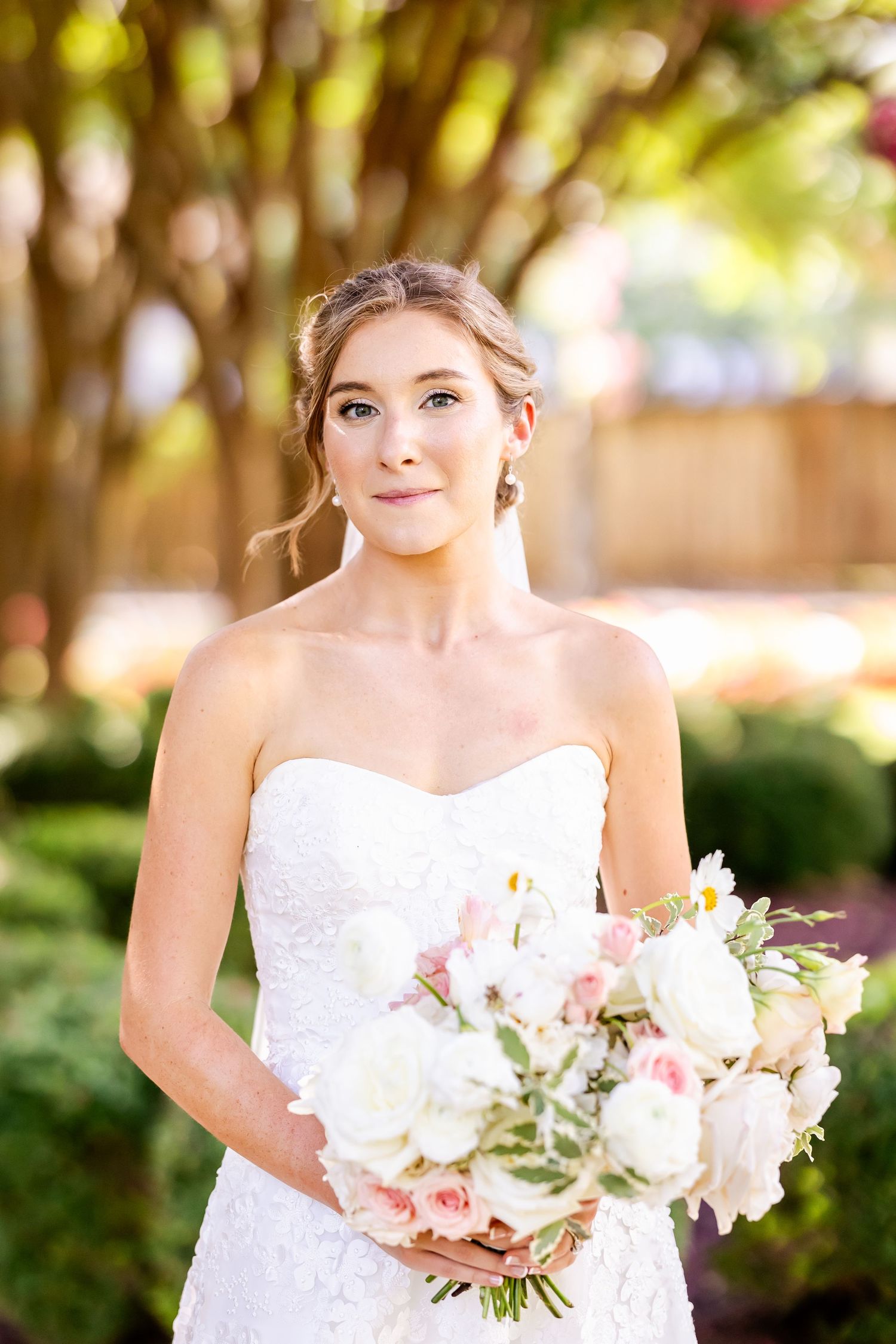 A bride in a white strapless lace gown holds a soft pink and white bouquet outdoors in a garden setting.