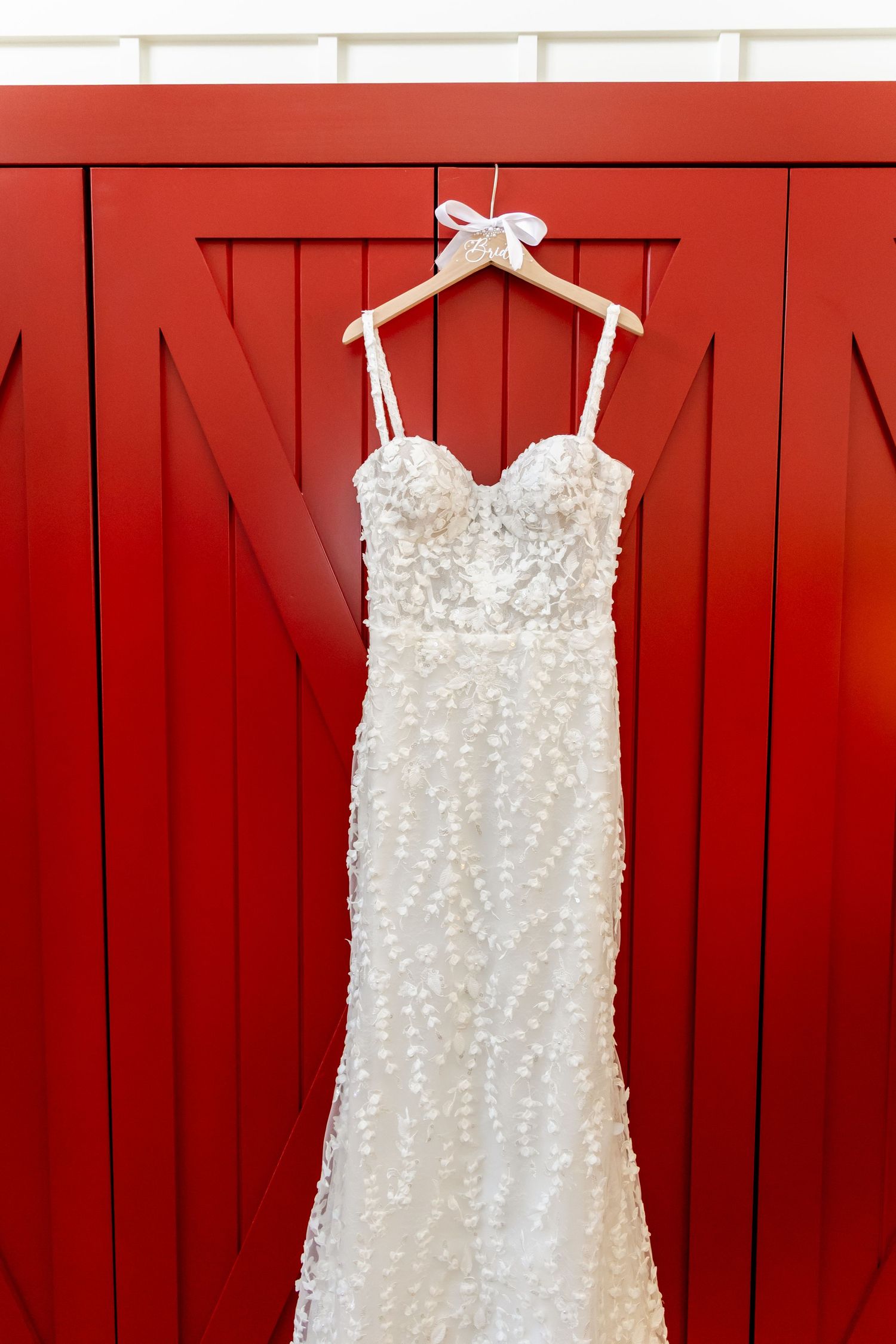 White beaded wedding dress hanging on wooden hanger against red barn door background.